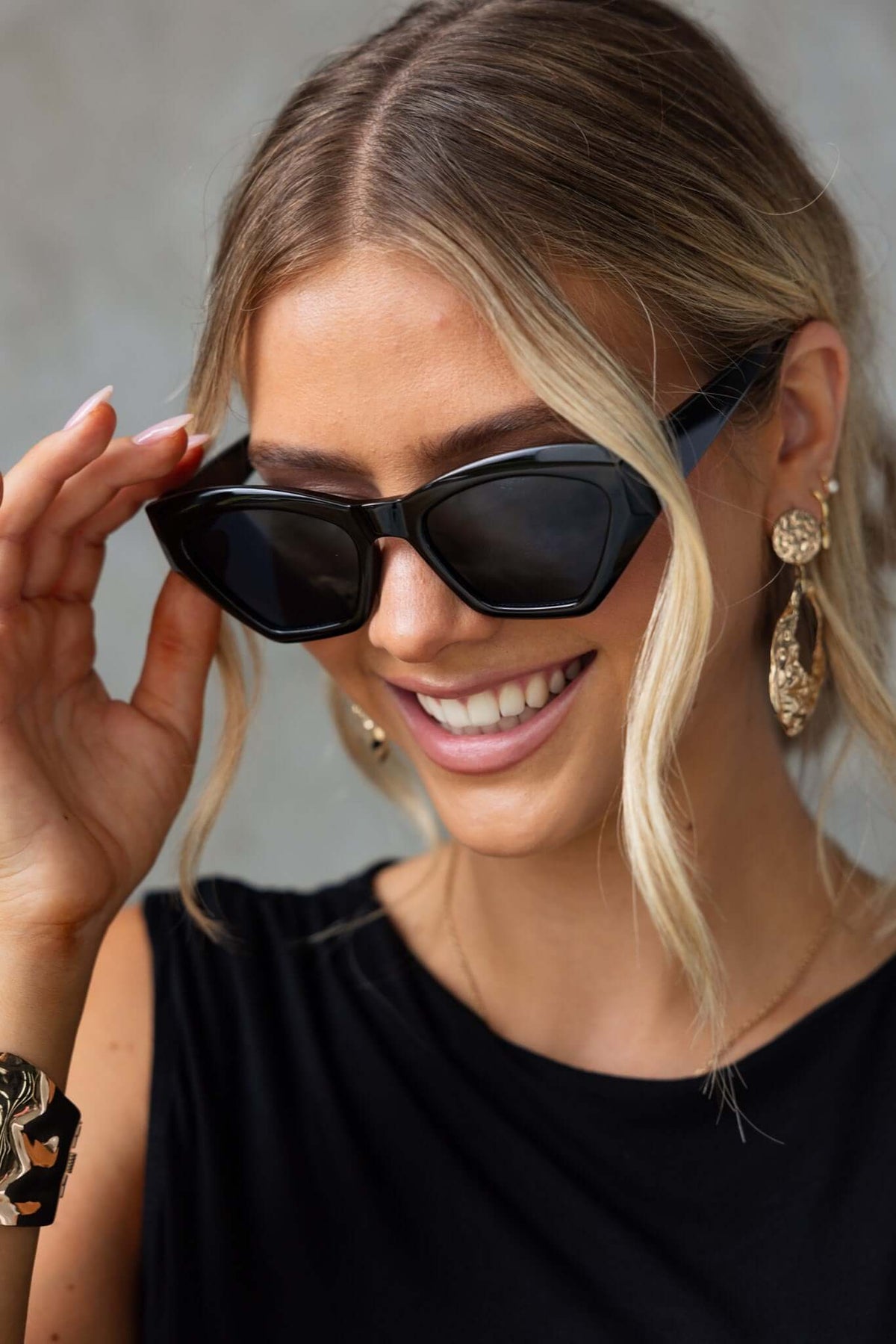 Woman wearing stylish black cat-eye sunglasses, smiling and showcasing trendy jewelry.