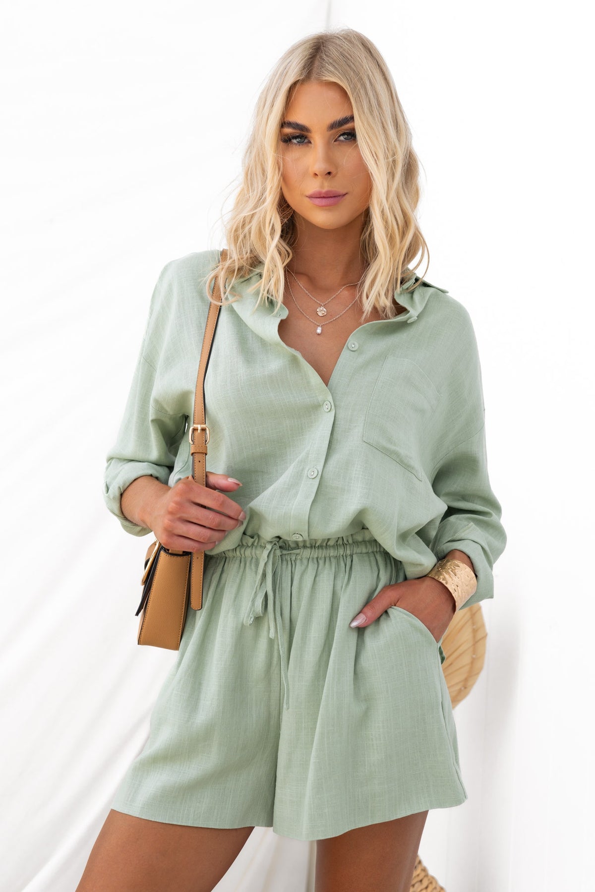 Model wearing Vanna Shorts in soft green with pockets and elastic waistband, styled with a button-up shirt and accessories.