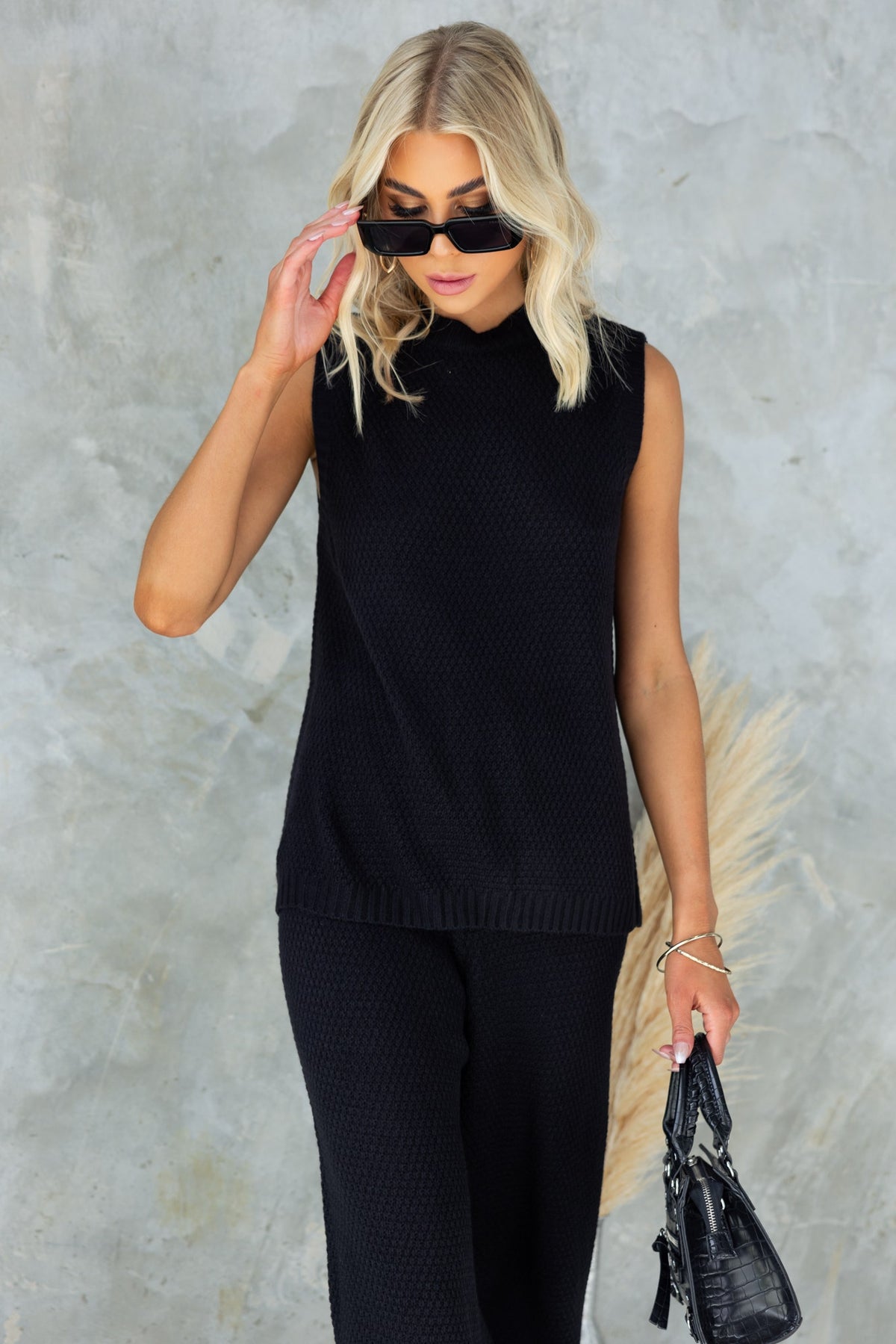 Model wearing Aera Top, a black sleeveless top with high neckline and open back, styled with sunglasses.