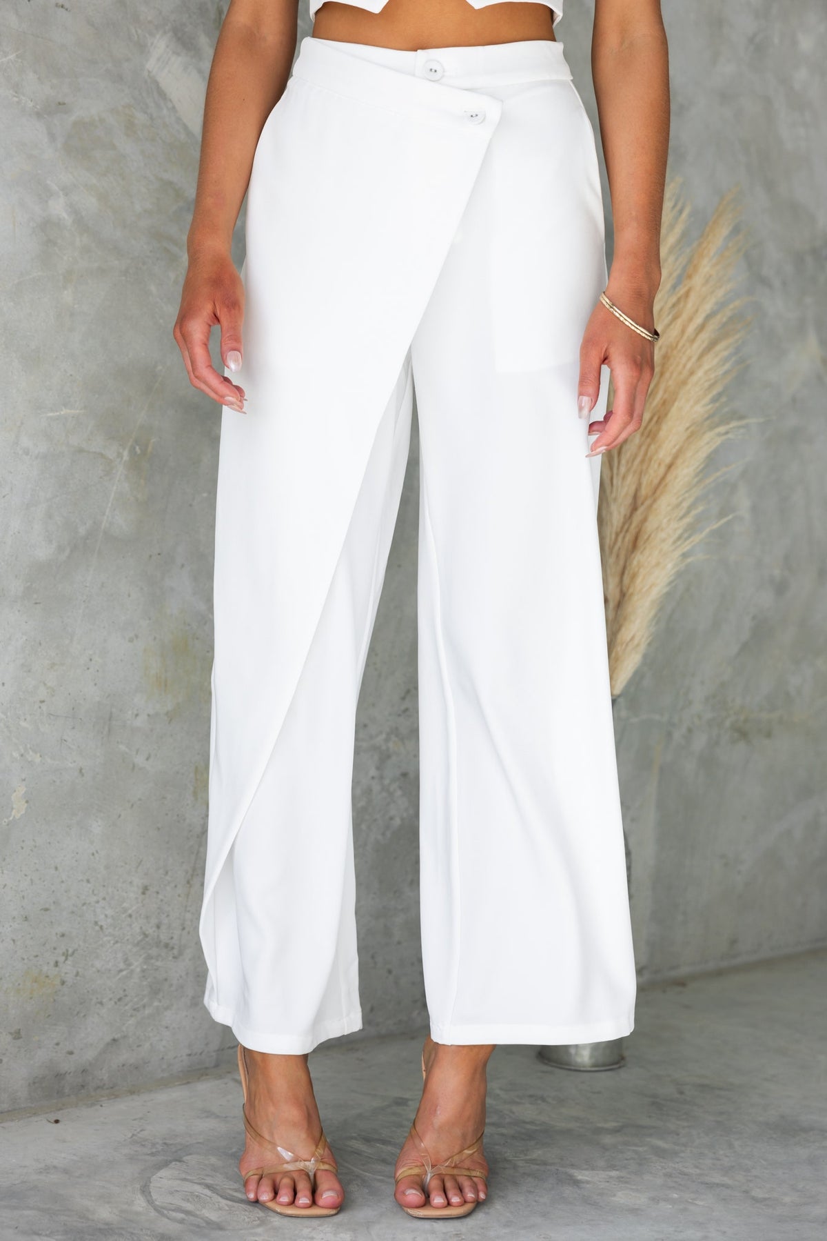 Elegant Oxford pants with asymmetric waist, mid-high fit, and pockets, styled with strappy heels in a modern setting.