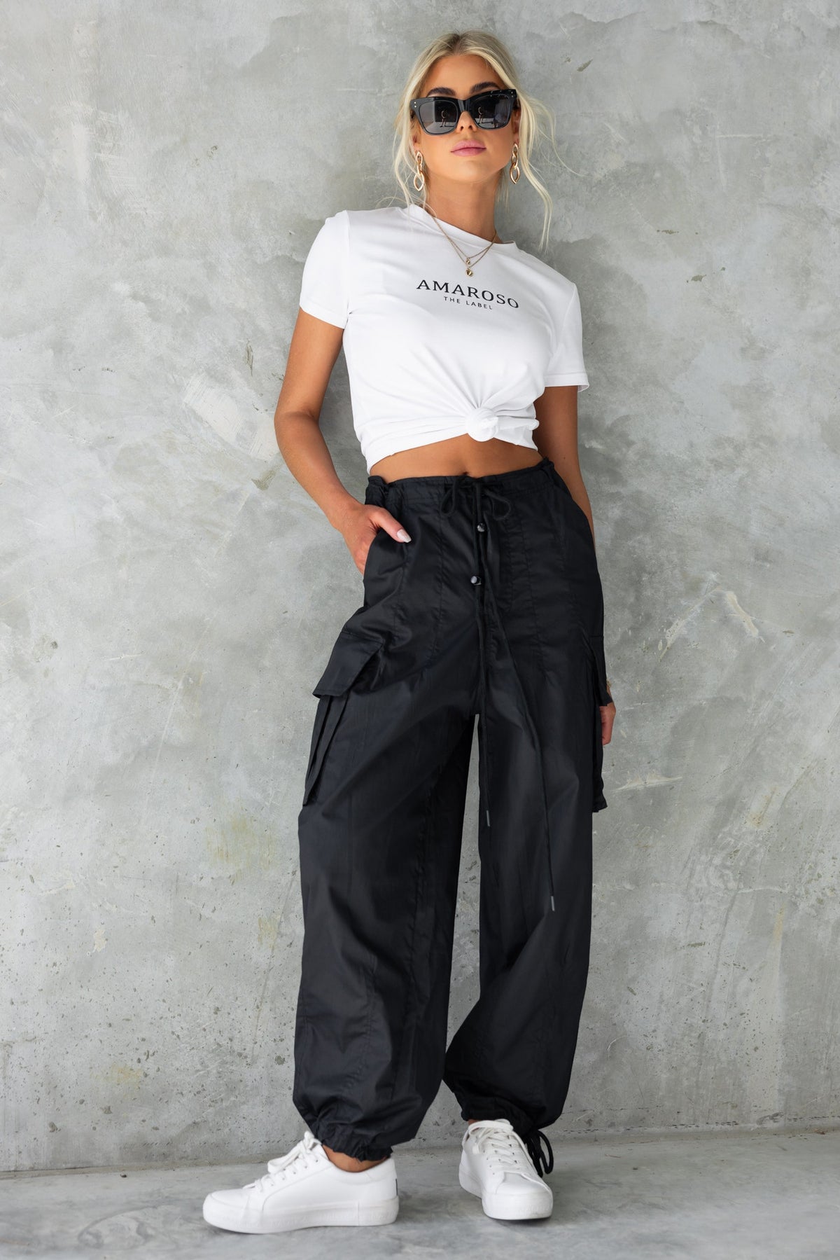 Model wearing black Conrad Cargo Pants with a white cropped shirt and sunglasses against a textured wall.