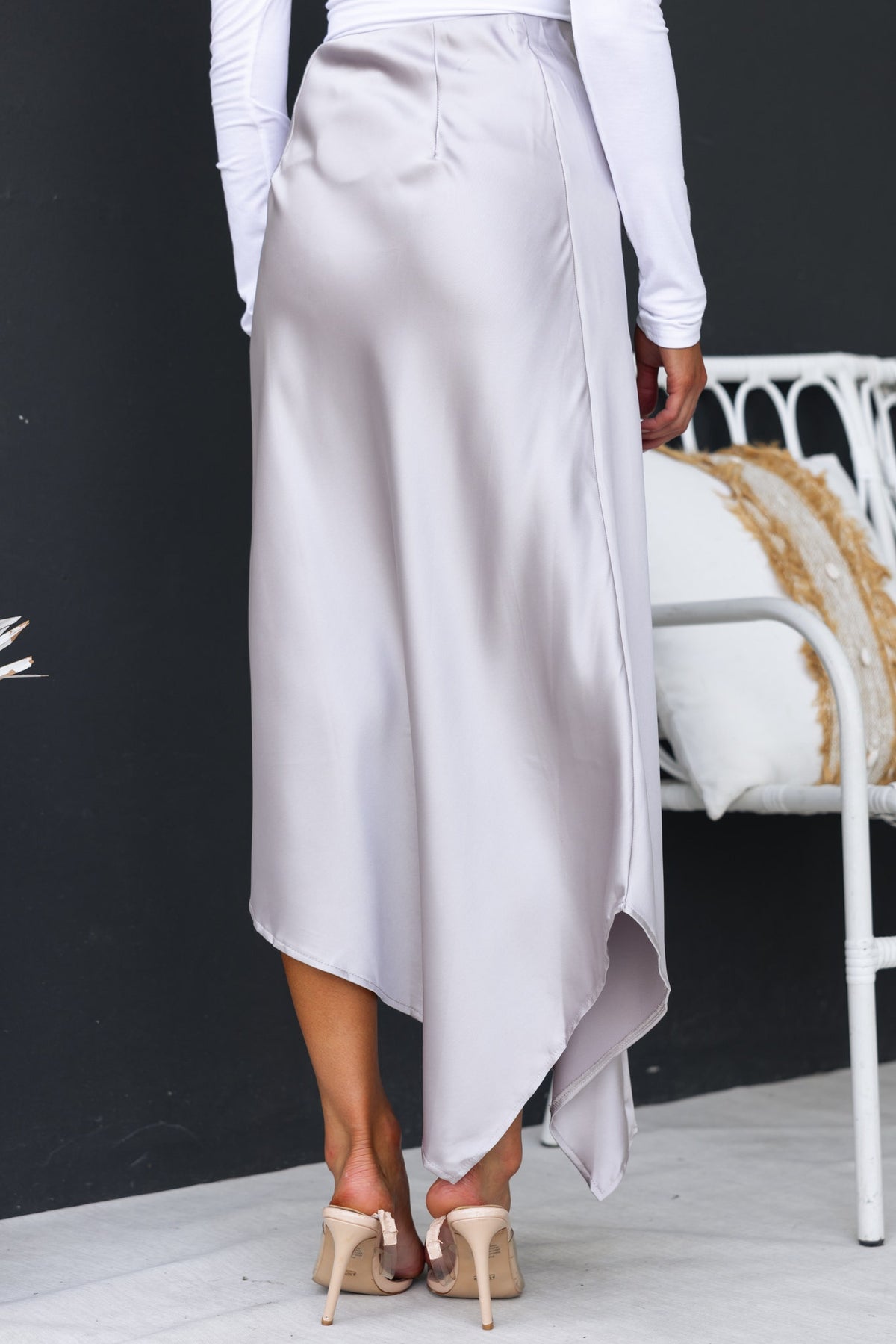 Back view of a Giallo Midi Skirt featuring a satin finish and an asymmetric hemline, styled with a white long-sleeve top.