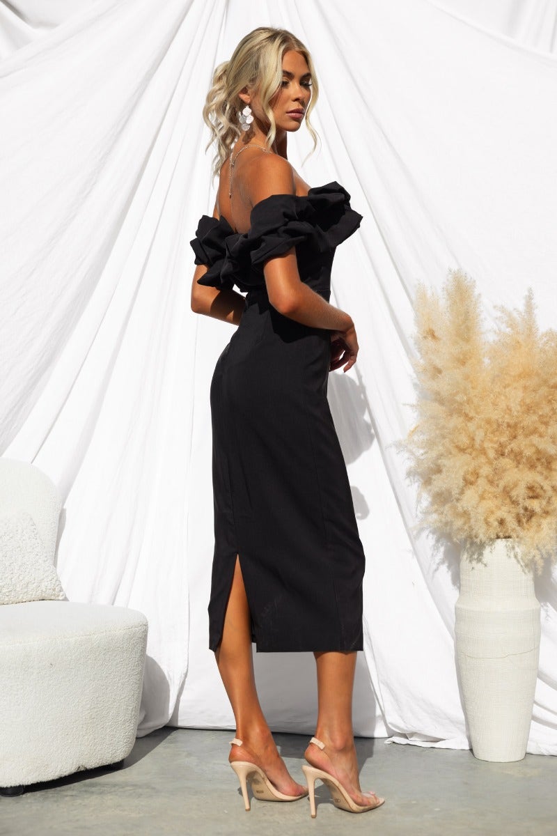 Elegant off-shoulder black midi dress with back split, model wearing heels, styled against a minimalist backdrop.