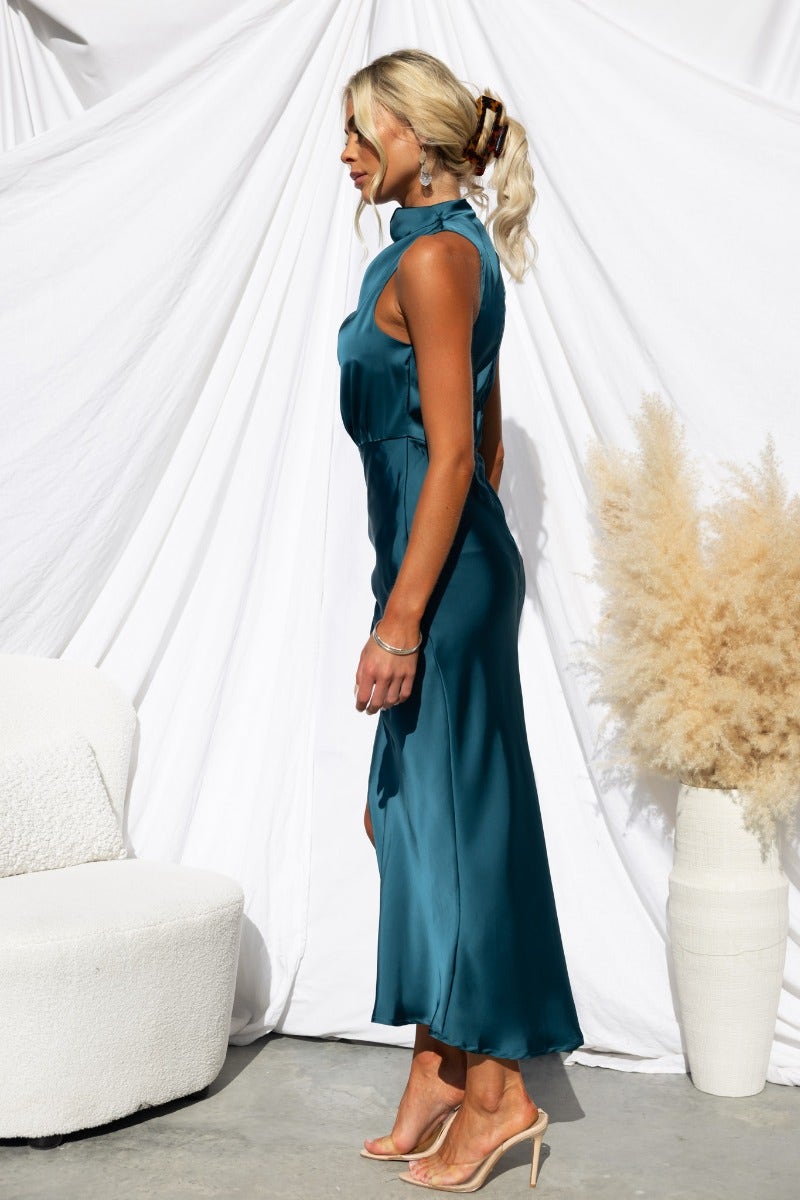 Model wearing a teal satin Honeymoon maxi dress with high neckline and asymmetric hemline, styled against a soft backdrop.