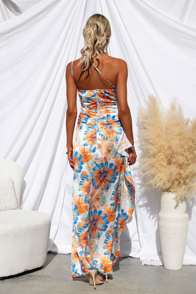 Back view of a floral satin maxi dress with one-shoulder neckline and ruffle hem, styled with natural surroundings.