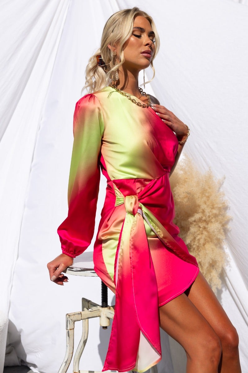 Model showcases the Tempest Dress, a vibrant one-shoulder satin dress with a wrap tie and long sleeves.
