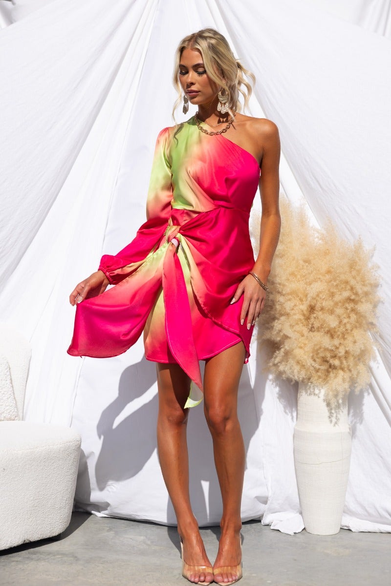 Model wearing a vibrant Tempest Dress with a one-shoulder neckline and wrap tie detail in pink and green hues.