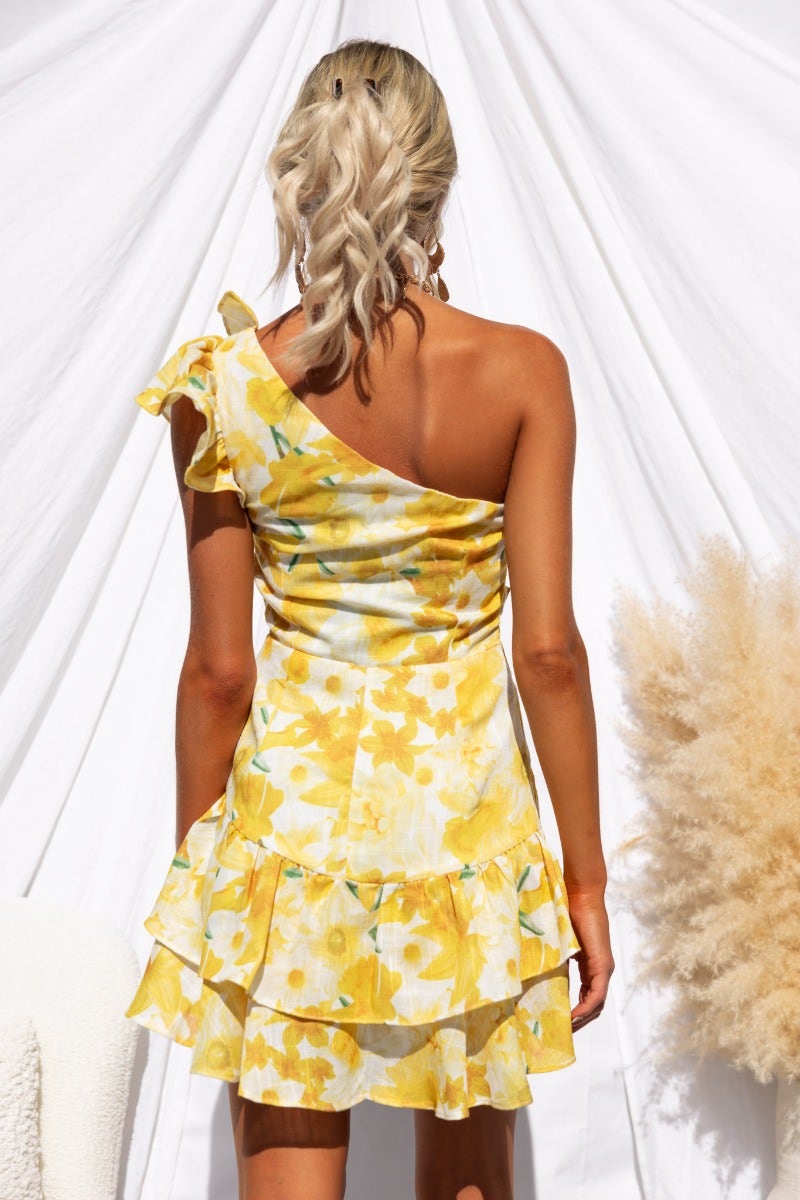 Model wearing a one-shoulder floral print dress in yellow with ruffle trim and layered skirt.