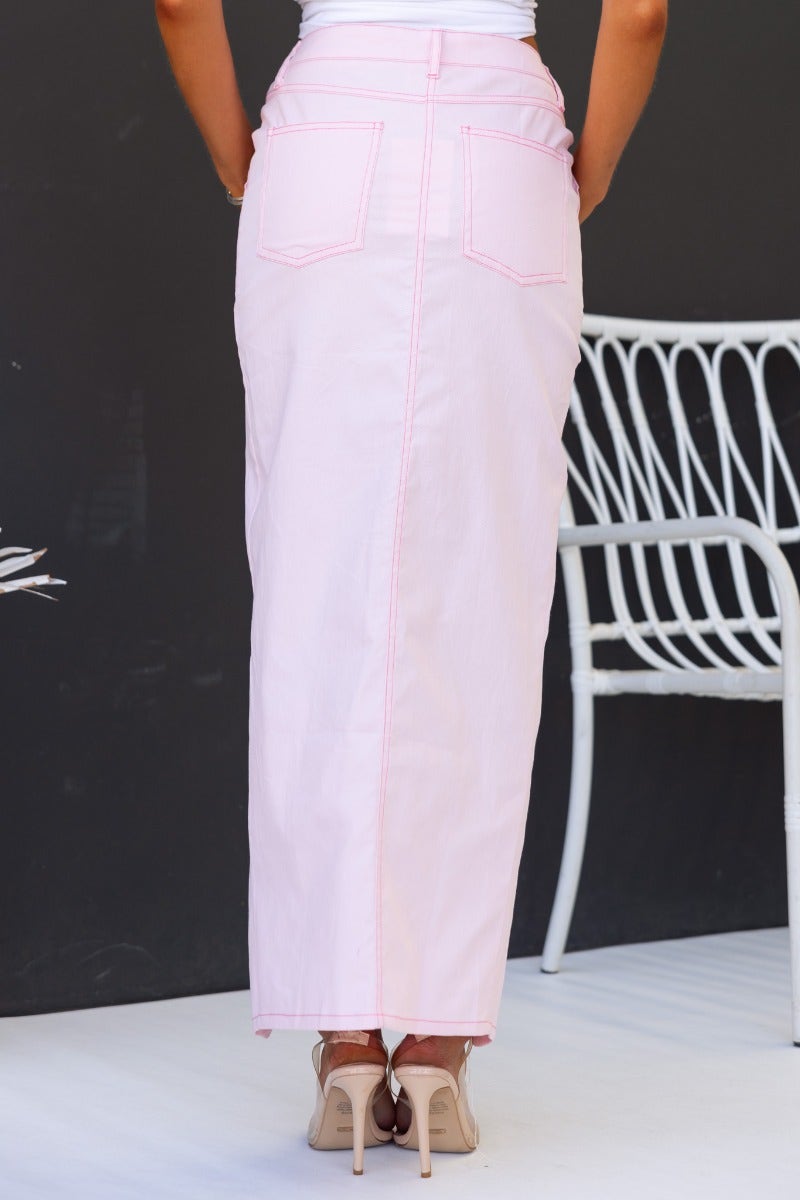 View of the back of a pink denim maxi skirt with pockets and high split, paired with stylish heels.