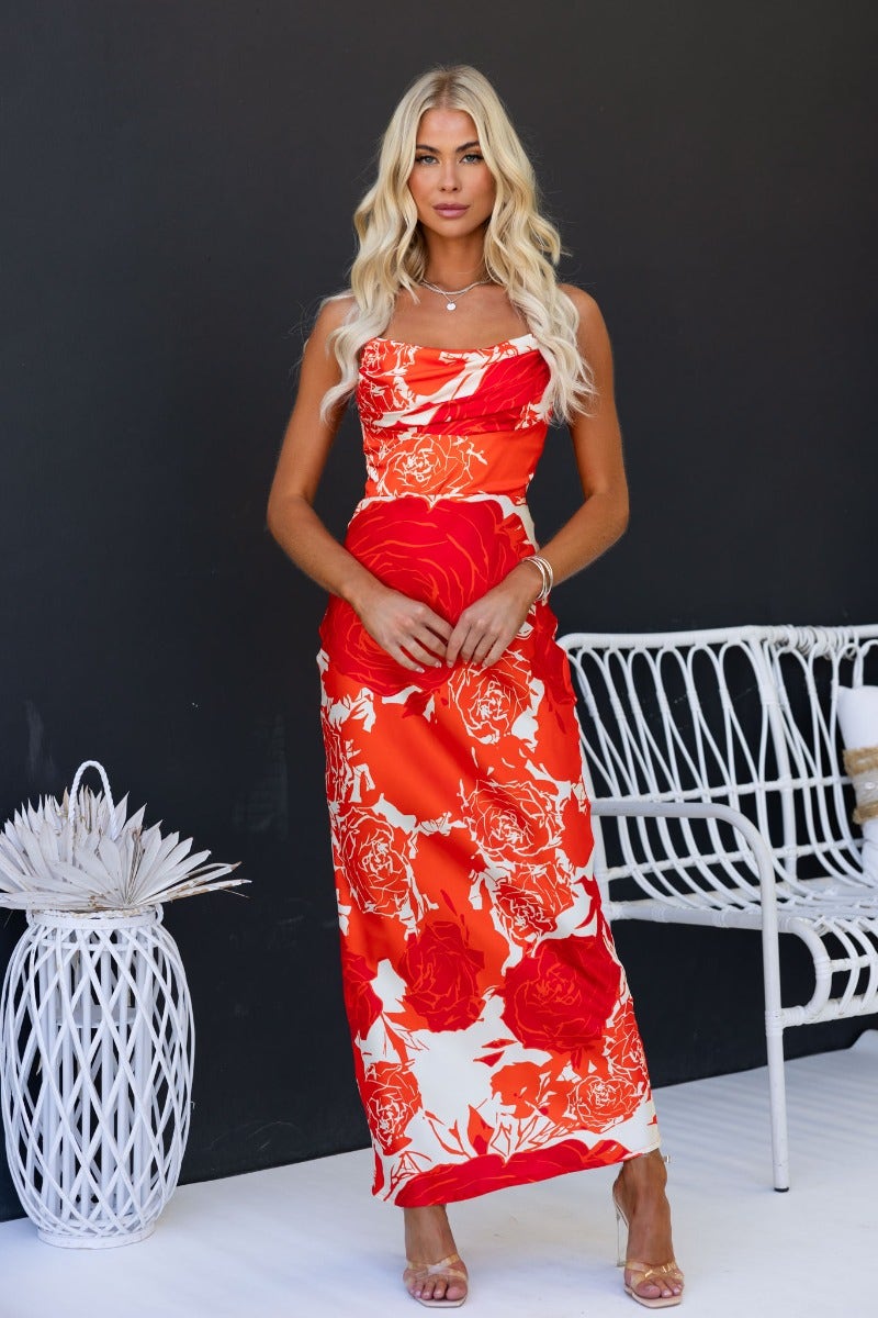 Model wearing Shana Maxi Dress with red floral design and cowl neckline against dark background.