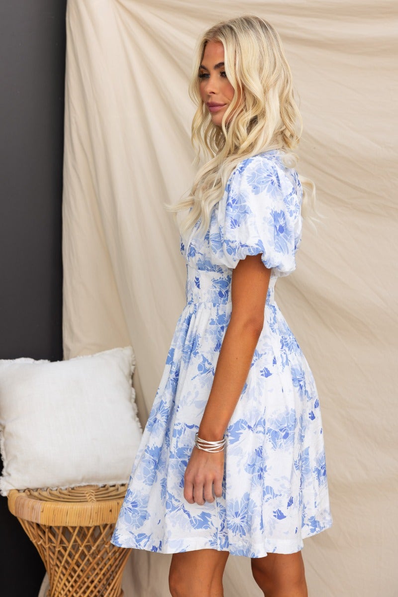 Floral print dress in blue and white, featuring puff sleeves and a fitted waist, styled with soft waves and a minimalist background.