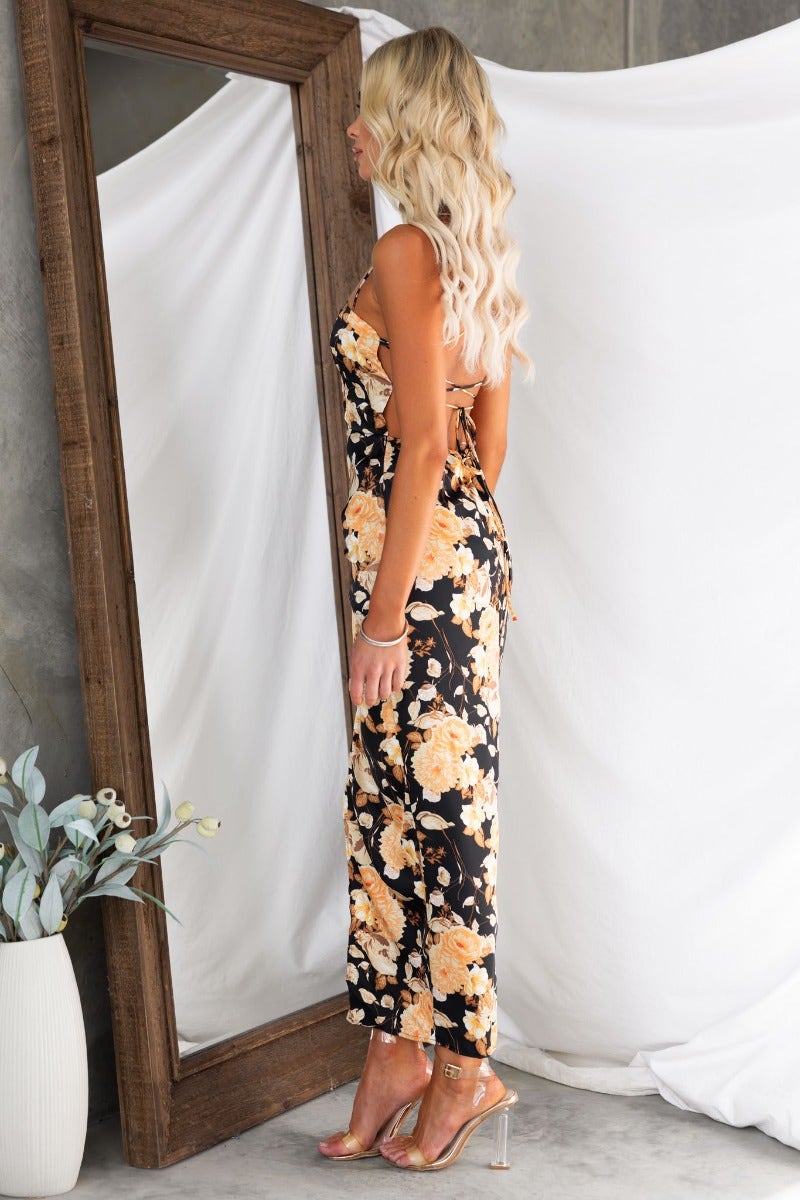 Model showcasing a floral black satin maxi dress with adjustable tie straps and a stylish cut-out back.