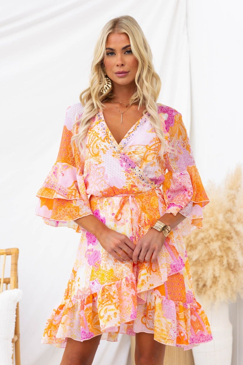 Model wearing a Fools Gold wrap dress with V-neckline, flared sleeves, and gem embellishments in vibrant orange and pink colors.