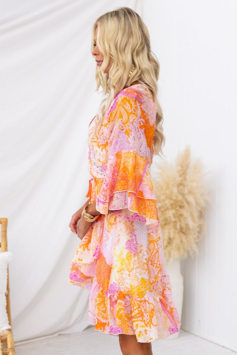 Model in colorful Fools Gold wrap dress with V-neckline and ruffled sleeves against a neutral background.