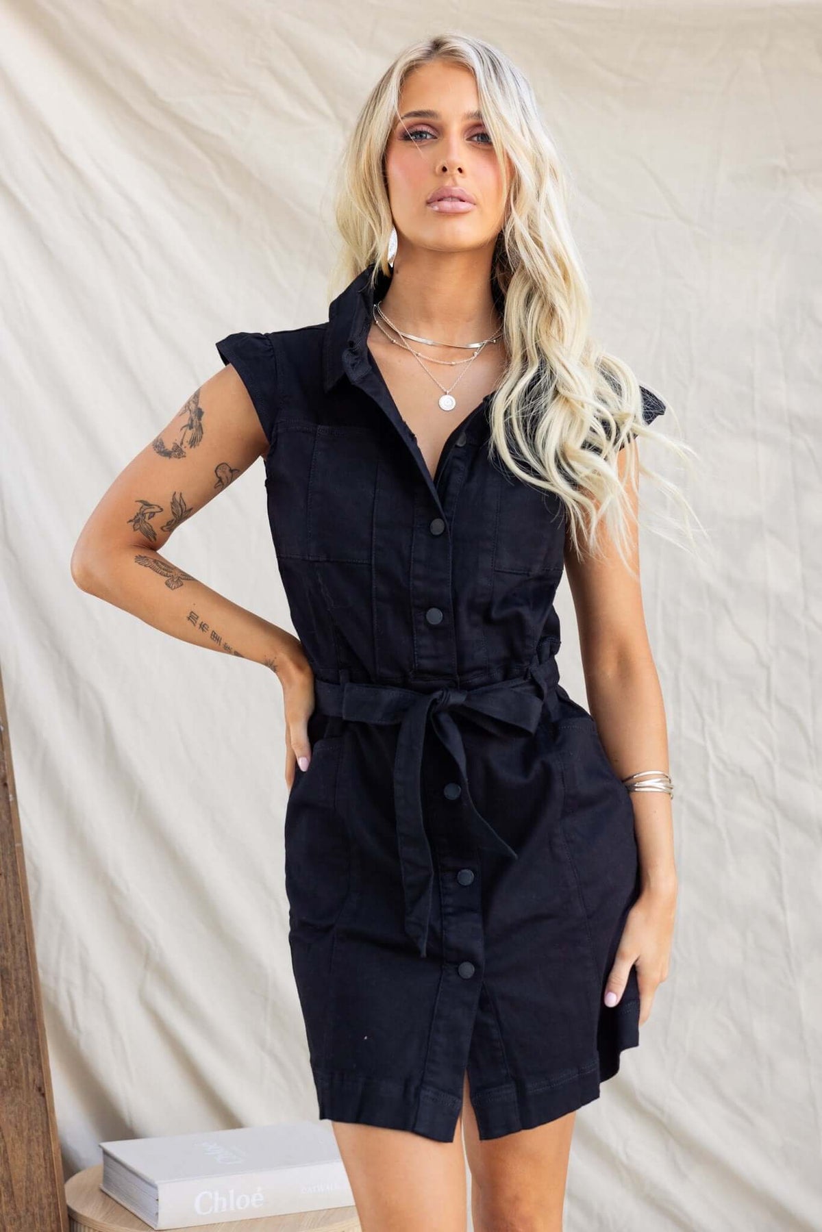 Model wearing a black denim dress with button-down front, removable belt, and pockets.