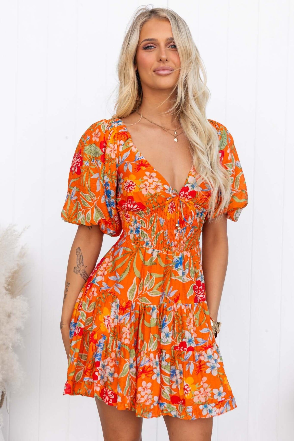 Airi Dress featuring a V-neckline, elastic sleeves, tie-back detail, and vibrant floral print in orange.