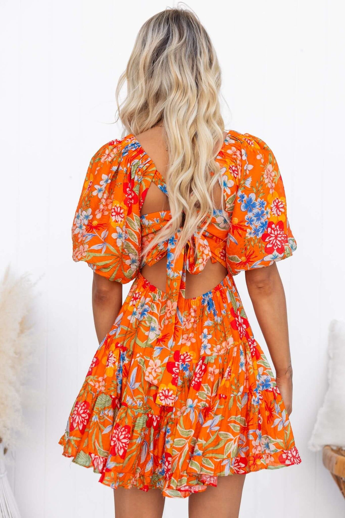 Back view of a woman in a vibrant orange floral Airi dress with a V-neckline and tie-back detail.