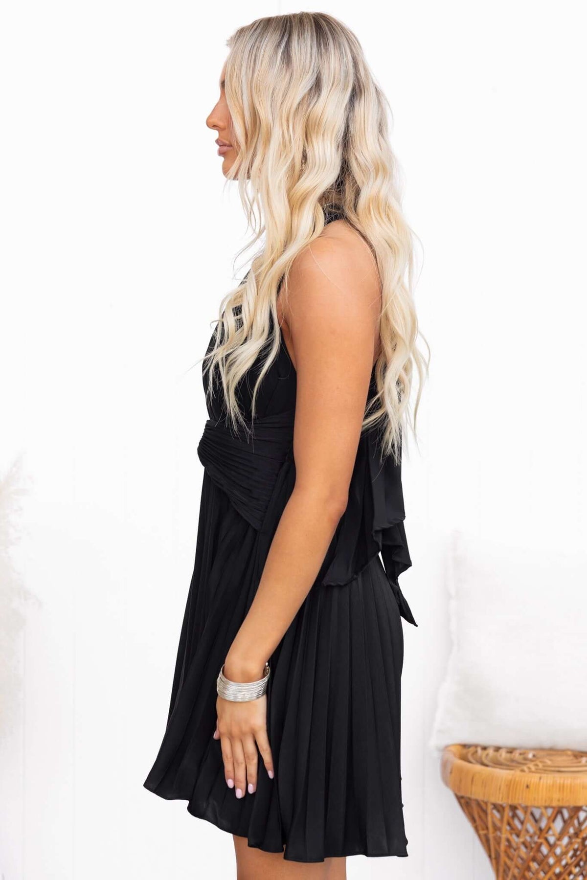 Side view of a woman wearing a black special day dress with adjustable tie neckline and pleated skirt.