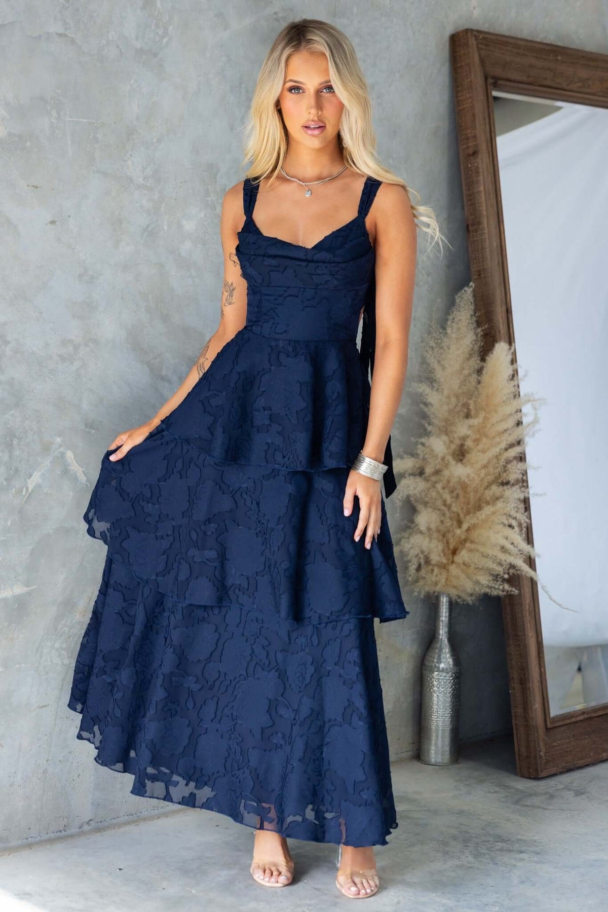 Model wearing a navy Rivoli maxi dress with tie straps, cowl neckline, and ruffle skirt, standing in front of a mirror.