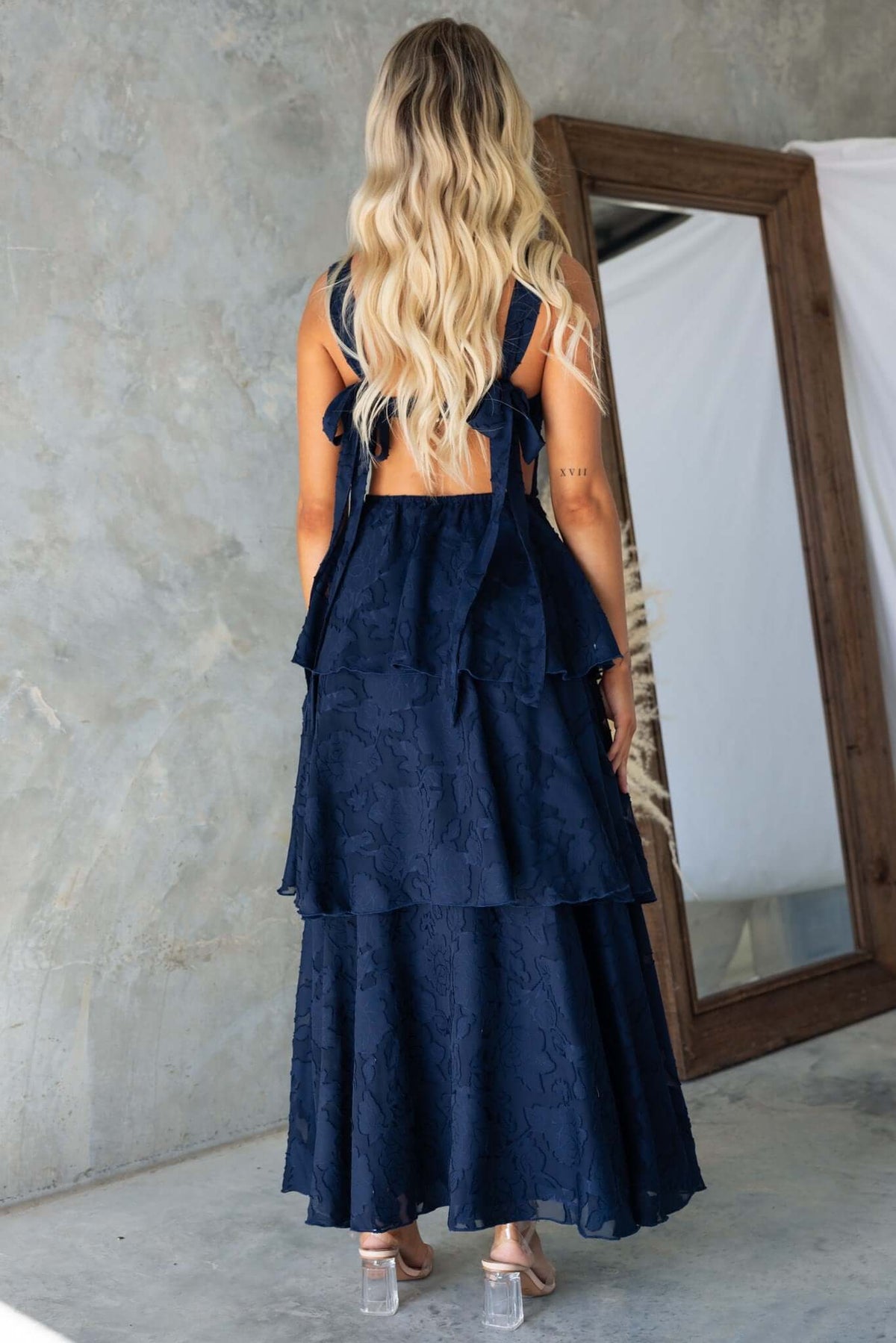 Back view of a woman wearing a navy Rivoli maxi dress with tie straps and ruffle skirt, standing near a mirror.