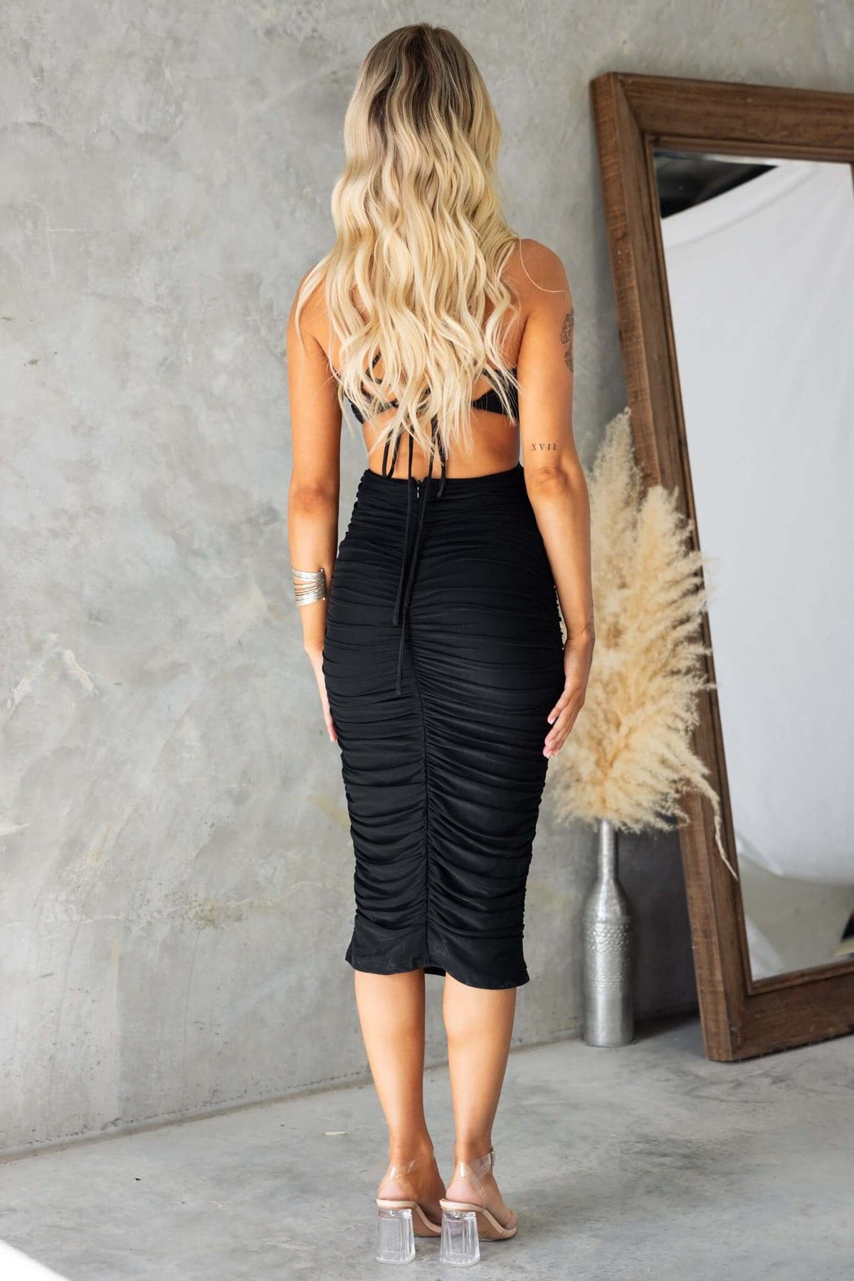 Back view of a woman wearing a black scarlet midi dress with adjustable tie straps and ruching details.
