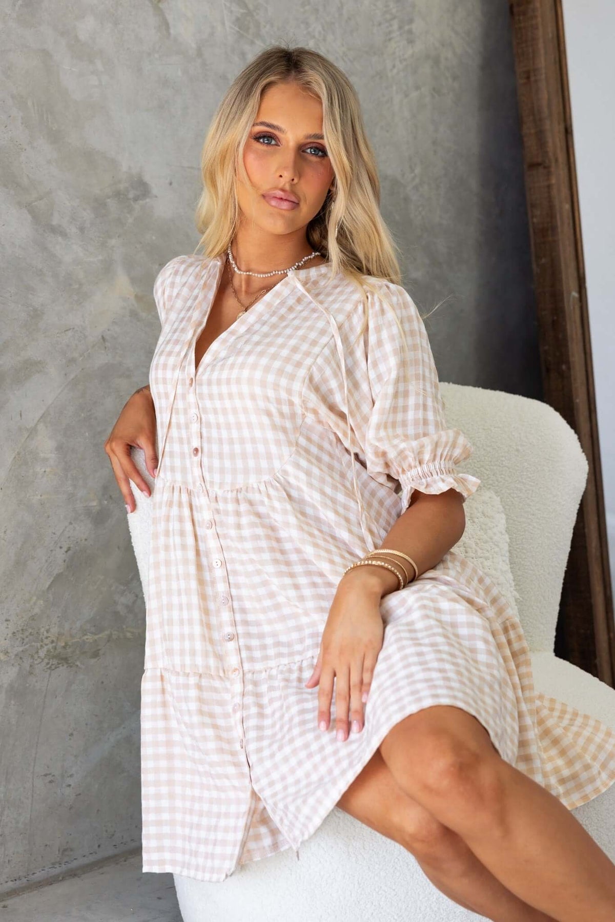 Model wearing Swinton Dress, an elastic-sleeve, button-up dress in a beige gingham pattern, styled casually.