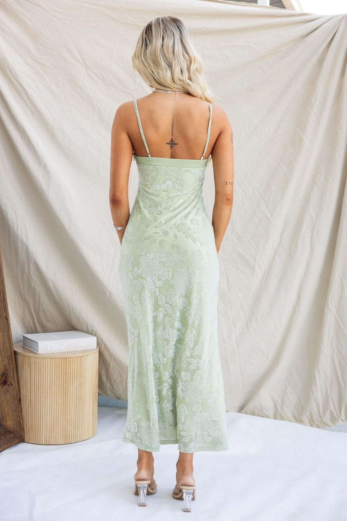 Back view of a woman in a green Melita maxi dress with adjustable straps and embossed velvet print, standing on a white surface.