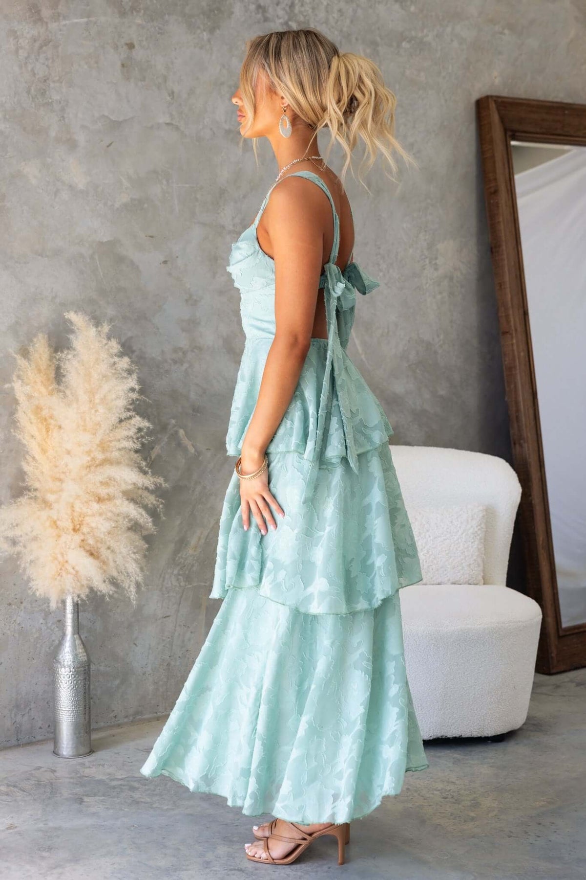 Model wearing a mint green Rivoli Maxi Dress with tie straps and layered ruffle skirt, displayed in a stylish interior.