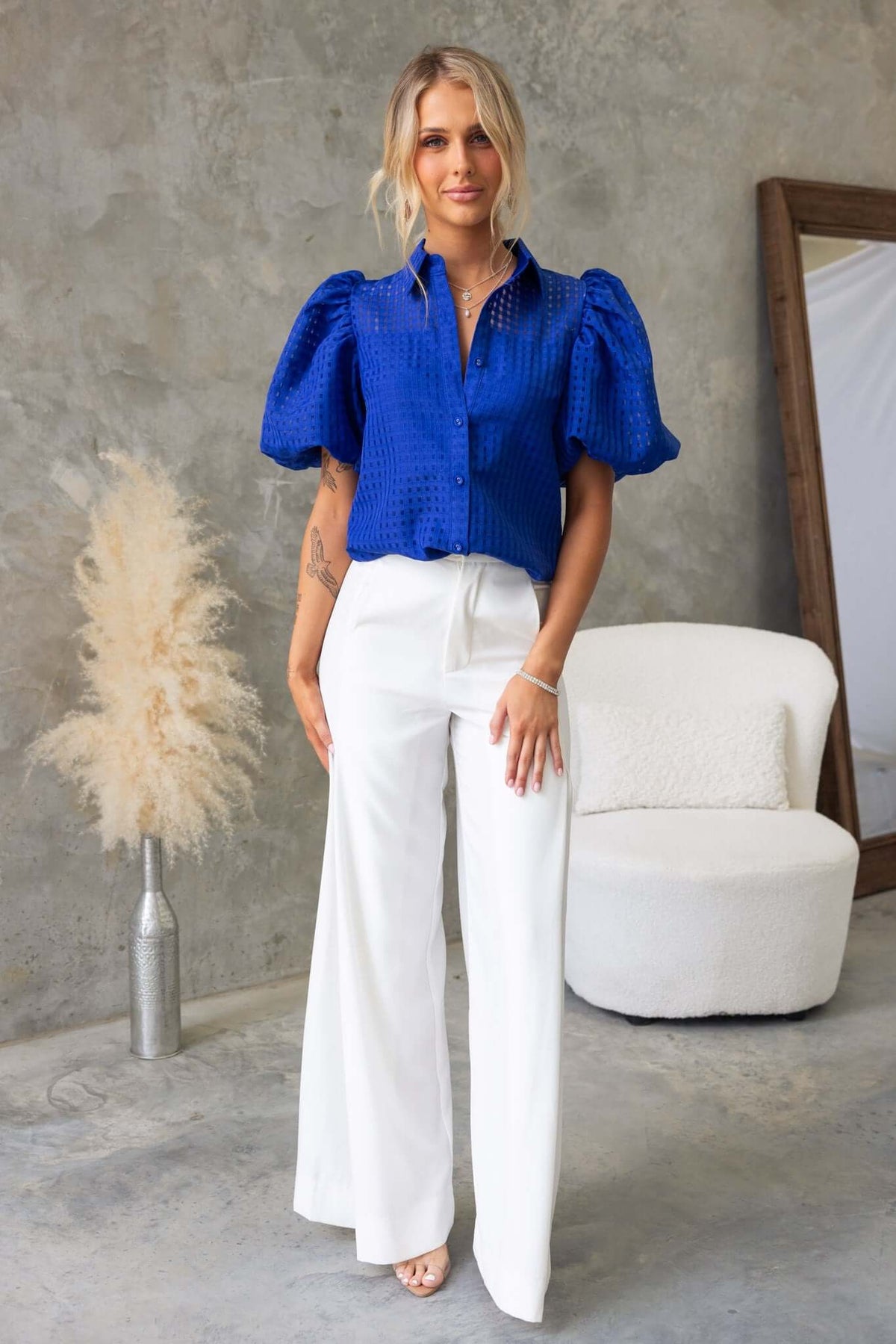 Model wearing Ursa Shirt in vibrant blue with puff sleeves, paired with white trousers in a stylish indoor setting.