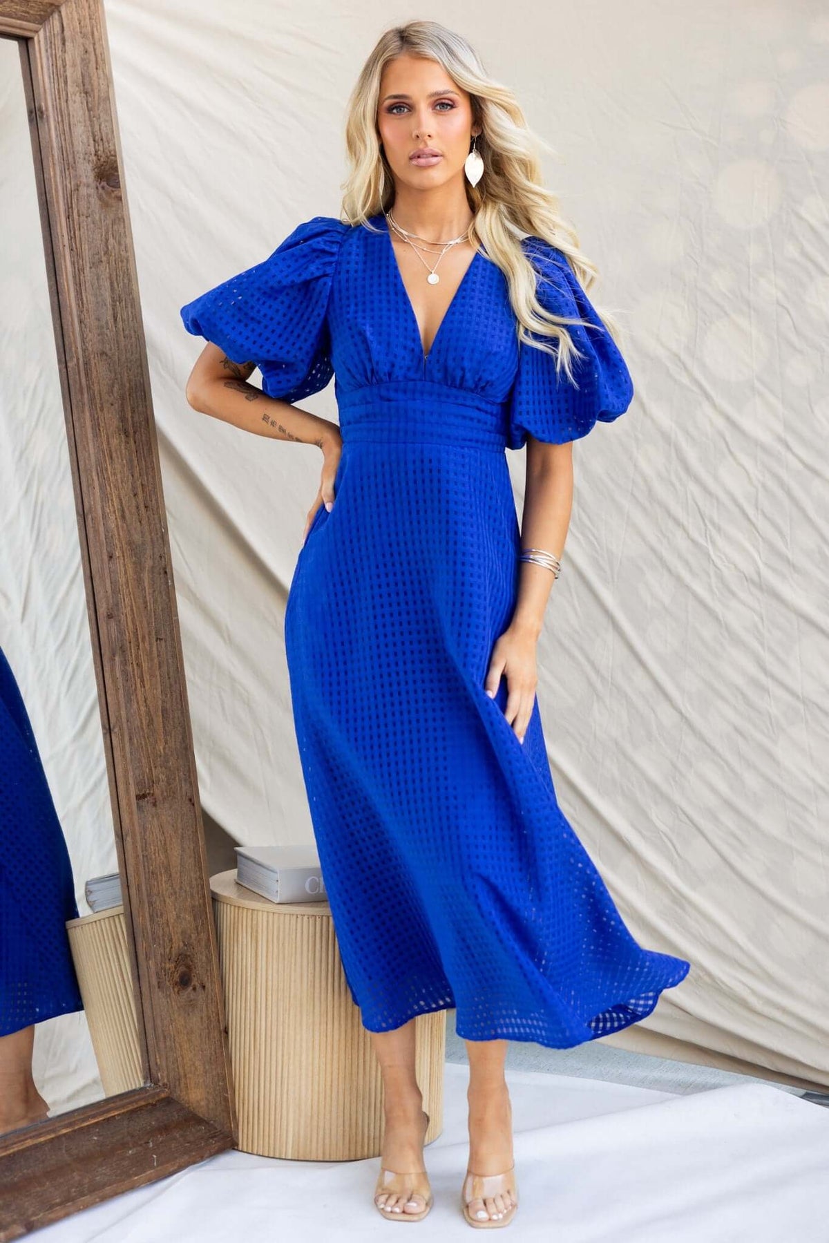 Model wearing a blue Nosita Midi Dress with V-neckline, puff sleeves, and pockets in a stylish indoor setting.