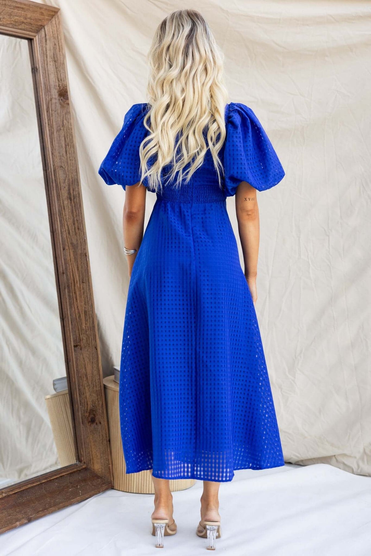 Back view of the Nosita Midi Dress, featuring a V-neckline, puff sleeves, and textured blue material.