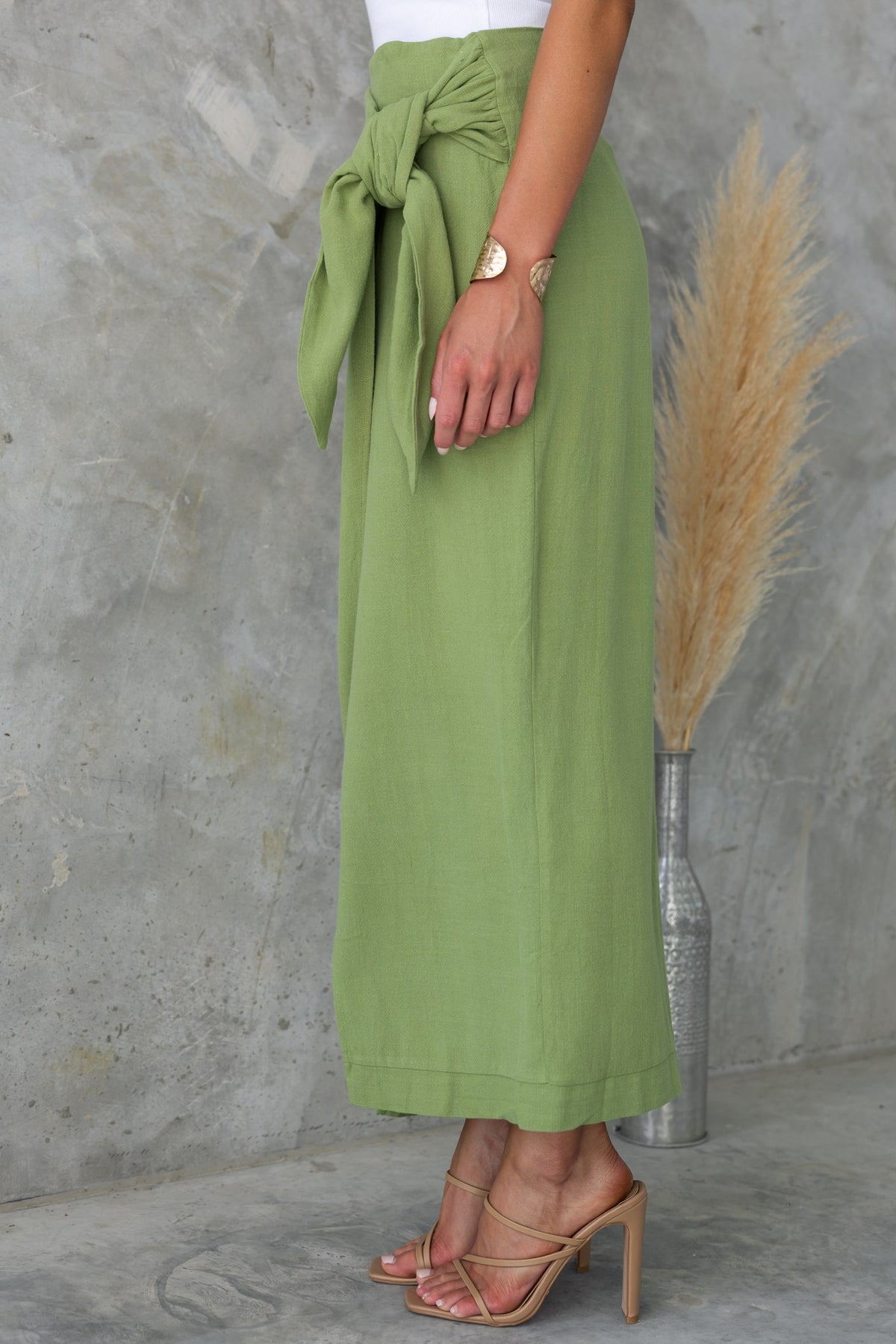 Side view of Tahnee Maxi Skirt in green, featuring a tie waist and asymmetric hemline, styled with elegant sandals.