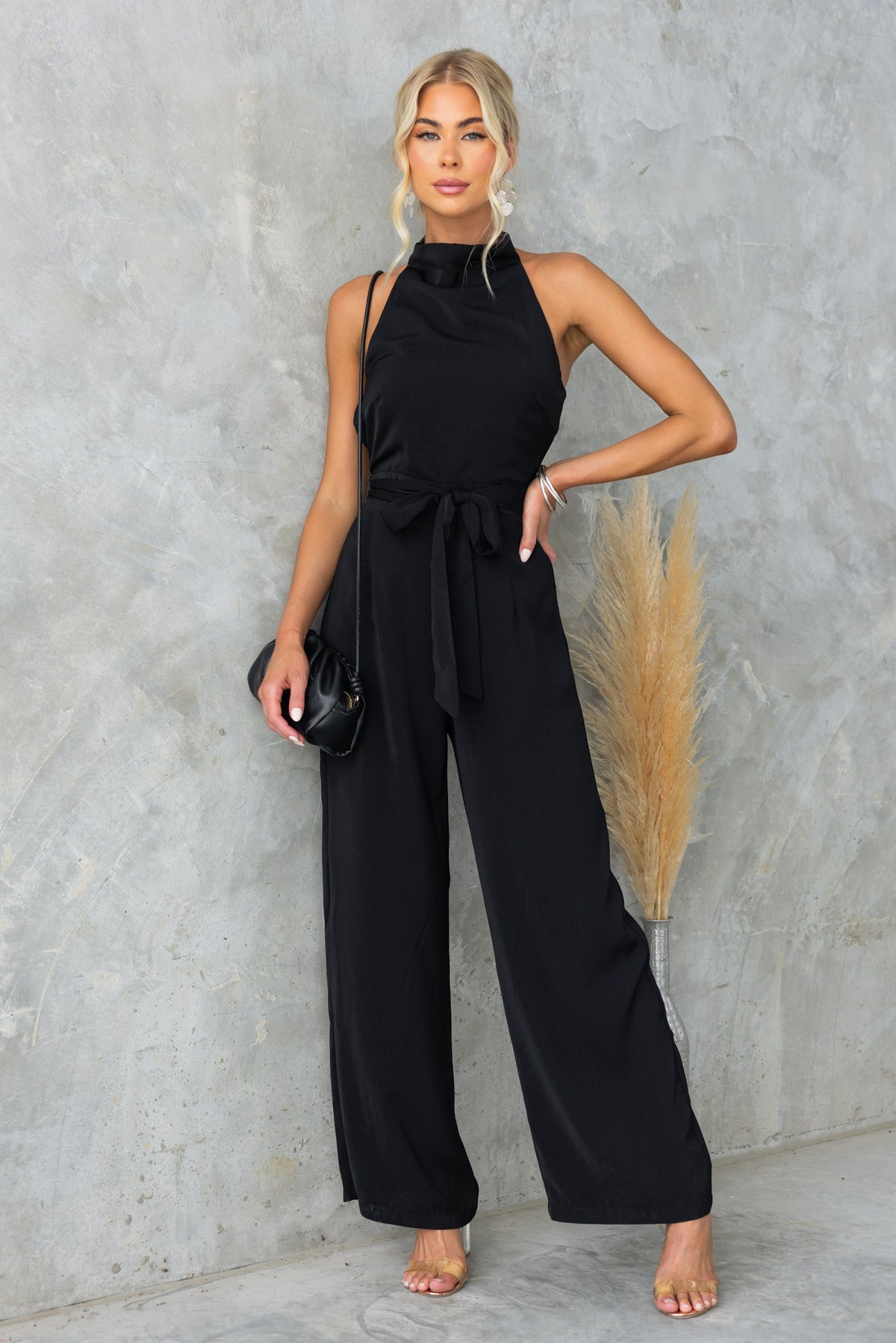 Model wearing a stylish black Skyler Jumpsuit with a high neckline and removable fabric tie against a textured background.