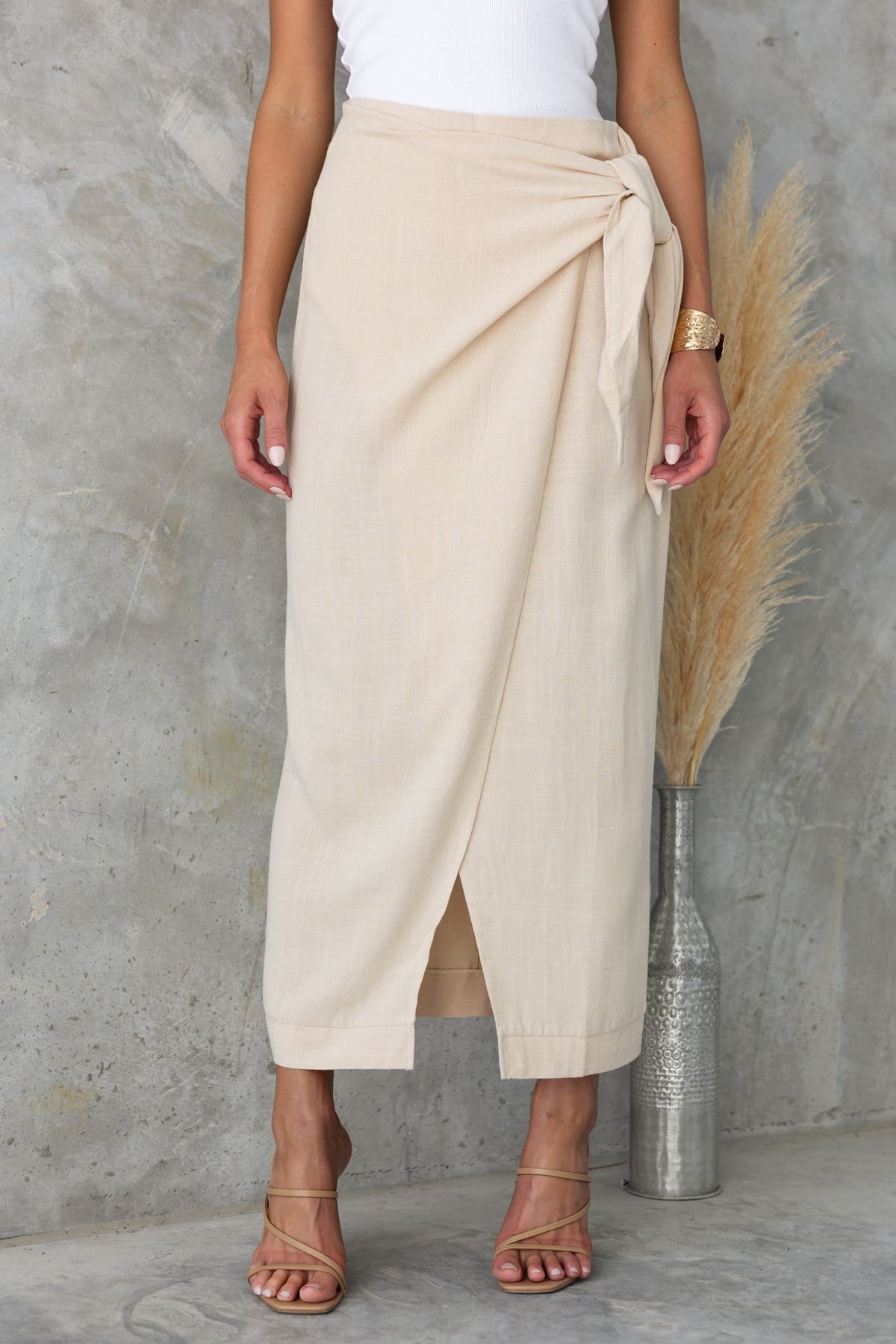 Model wearing Tahnee Maxi Skirt in beige with a wrap design, side tie, and asymmetric hem against a neutral background.