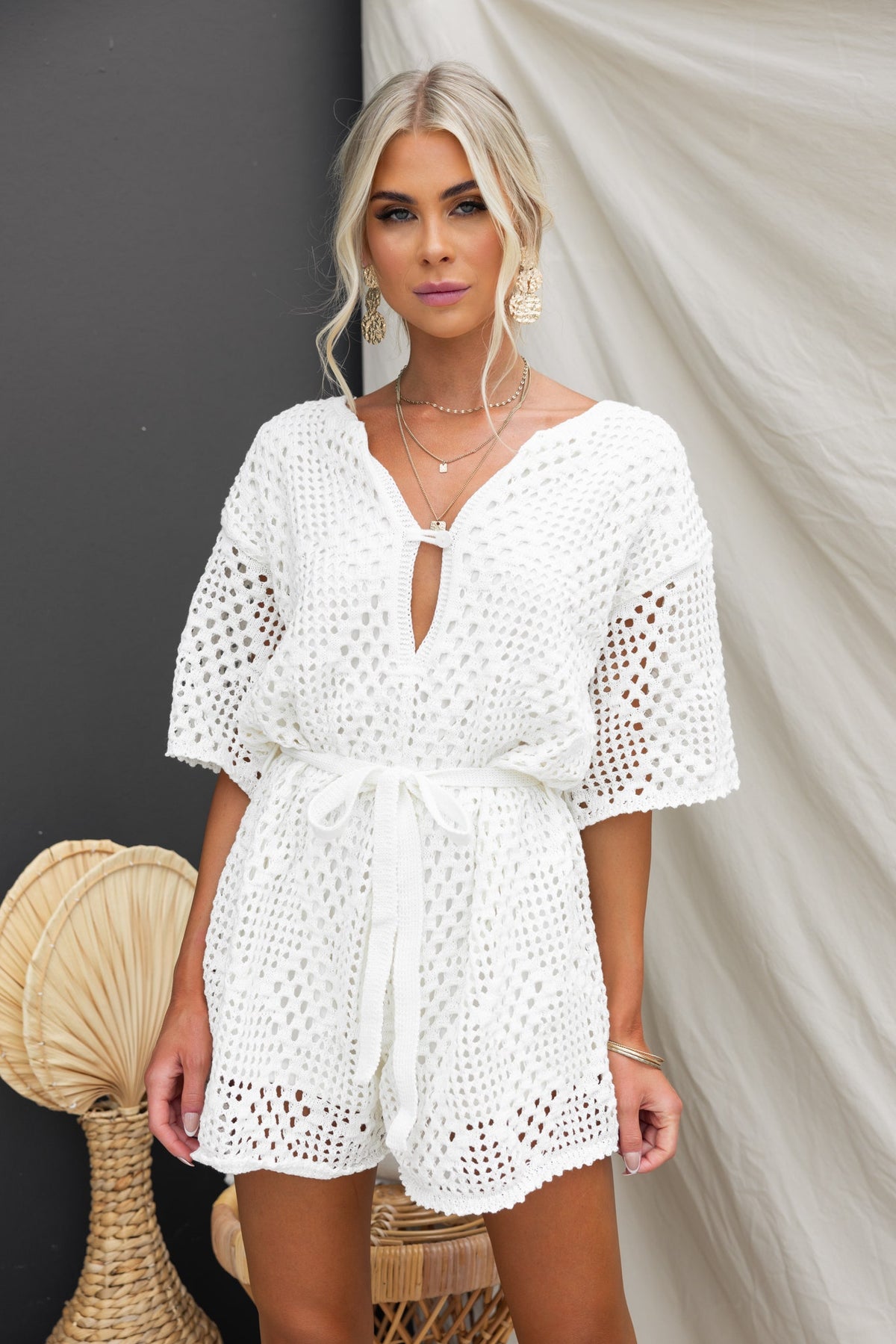 Model in a white crochet Momo Playsuit featuring a button detail and removable tie, styled with layered jewelry.