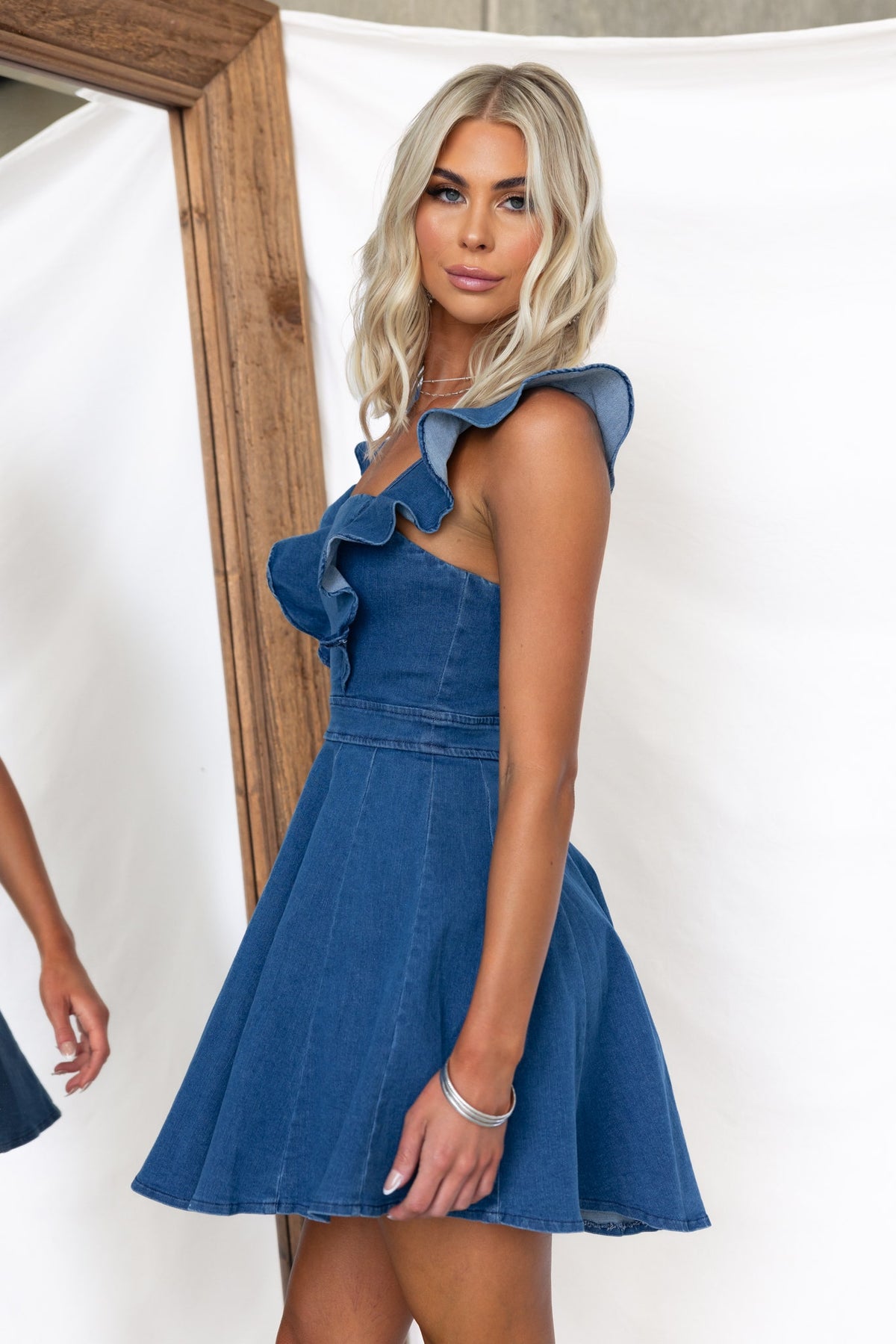 Model wearing a Natty Denim Dress with ruffled straps, showcasing a fitted bust and flared skirt against a minimalist backdrop.