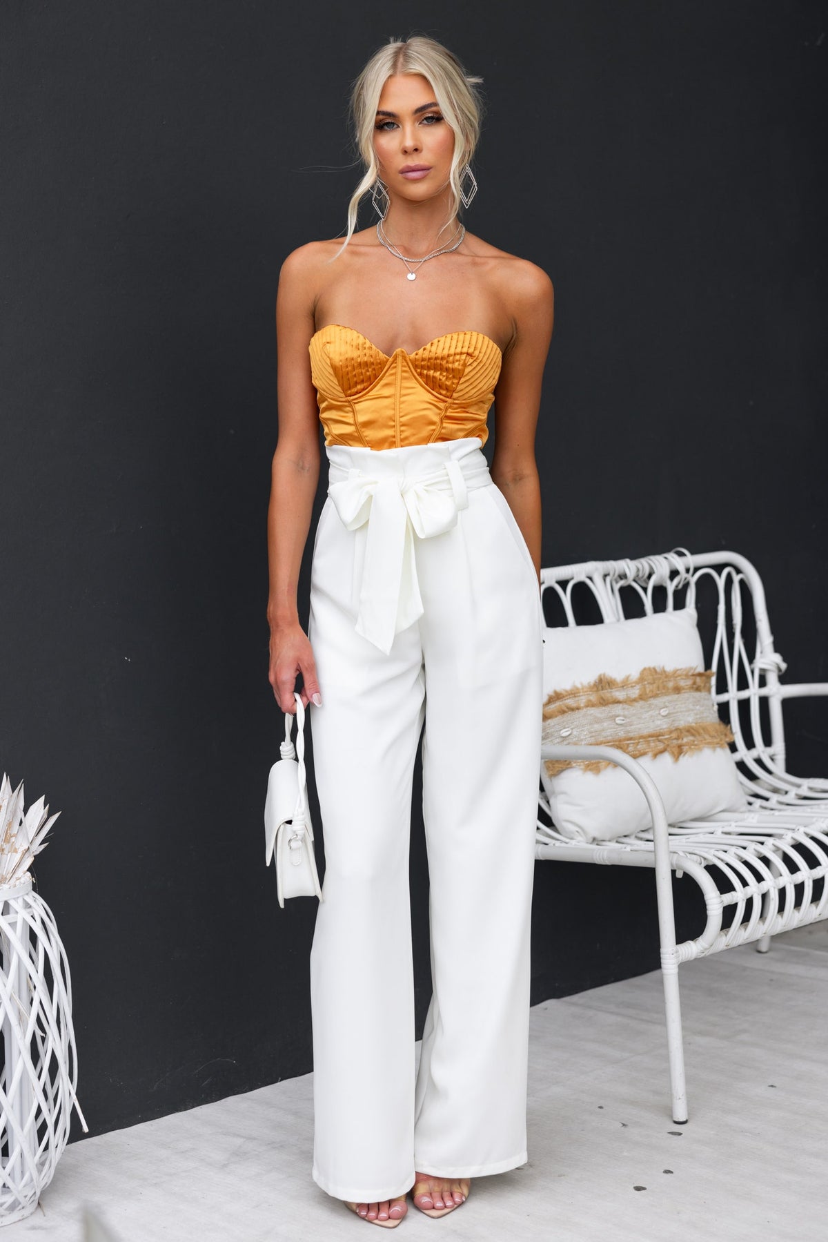 Model wearing a strapless Priya Top in orange with high-waisted white pants, posing against a dark wall.