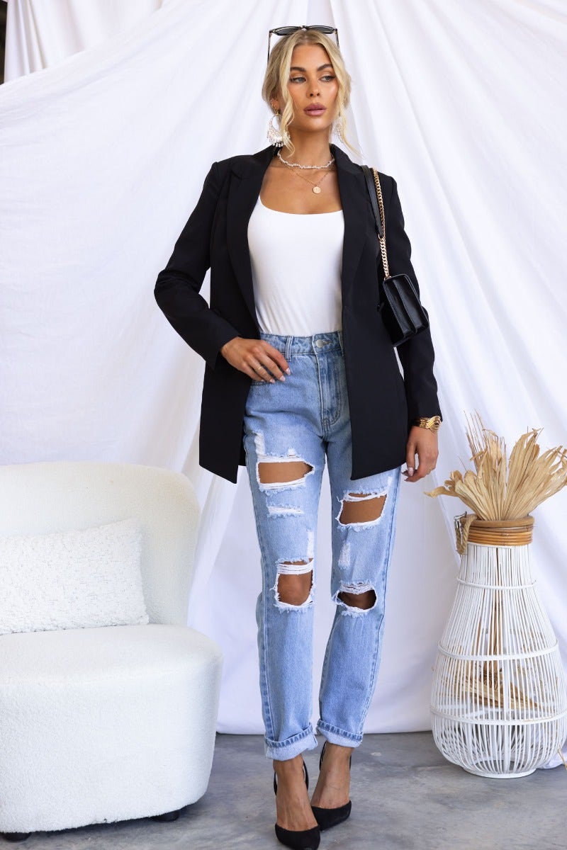 Stylish woman in a black Zeva blazer, white top, and ripped jeans, showcasing a trendy look with a modern backdrop.