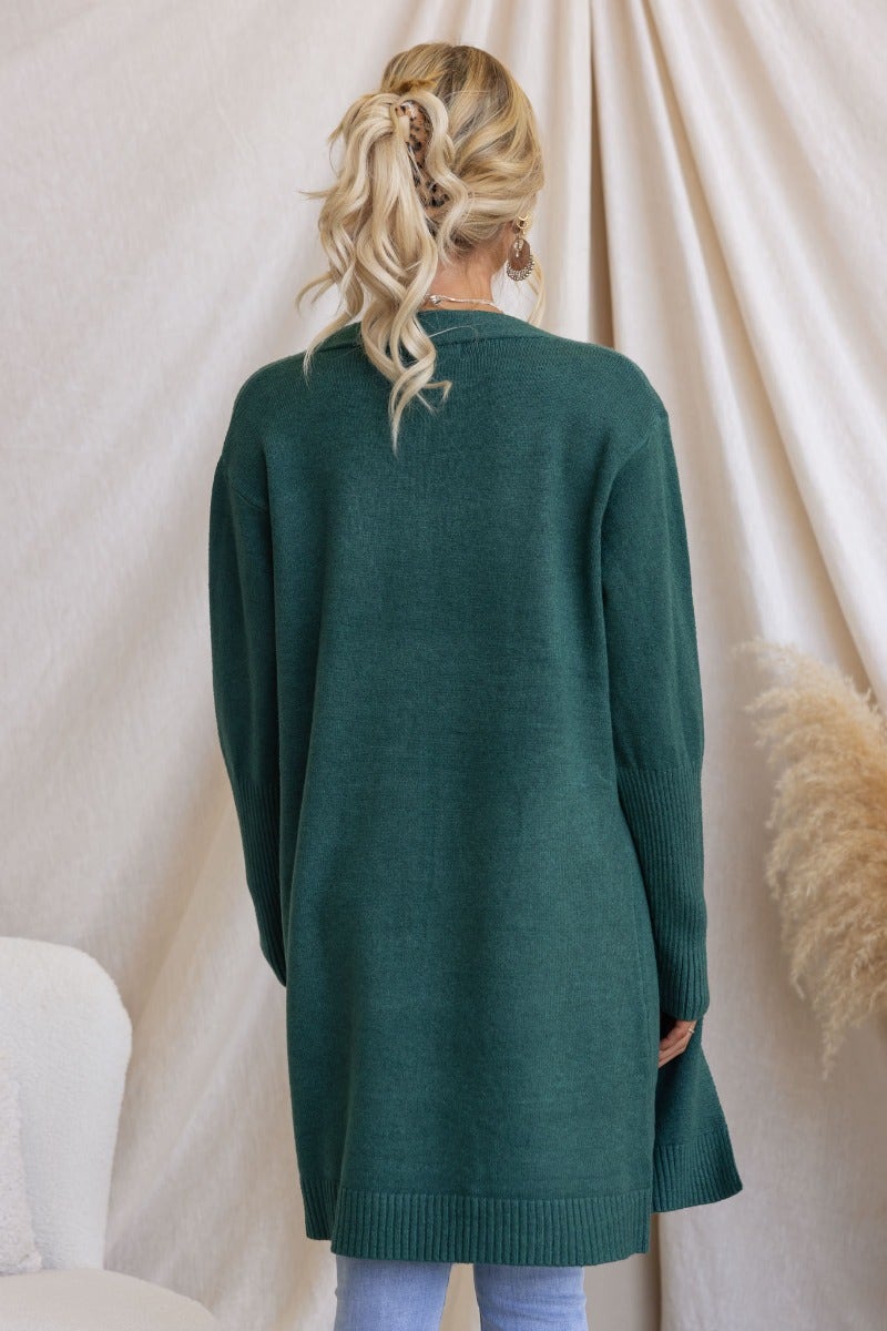 Back view of a woman wearing a longline green ribbed knit cardigan with pockets, styled with casual jeans.