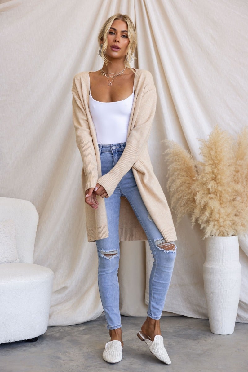 Stylish woman in a ribbed knit longline cardigan, paired with ripped jeans and mules, against a soft backdrop.
