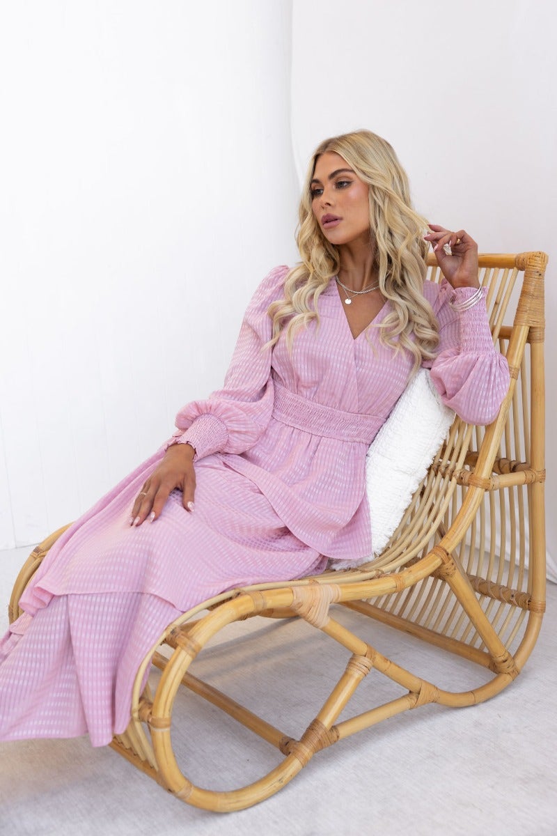 Model wearing Briena Midi Dress in pink, sitting on a rattan chair, showcasing the dress's elegant design and ruffle details.