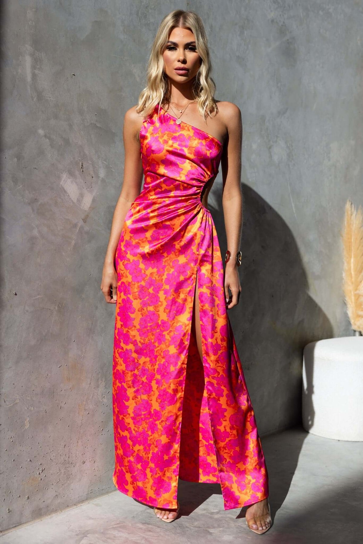 One-shoulder floral maxi dress in pink satin with high split and elastic cut-out detail, modeled by a woman.