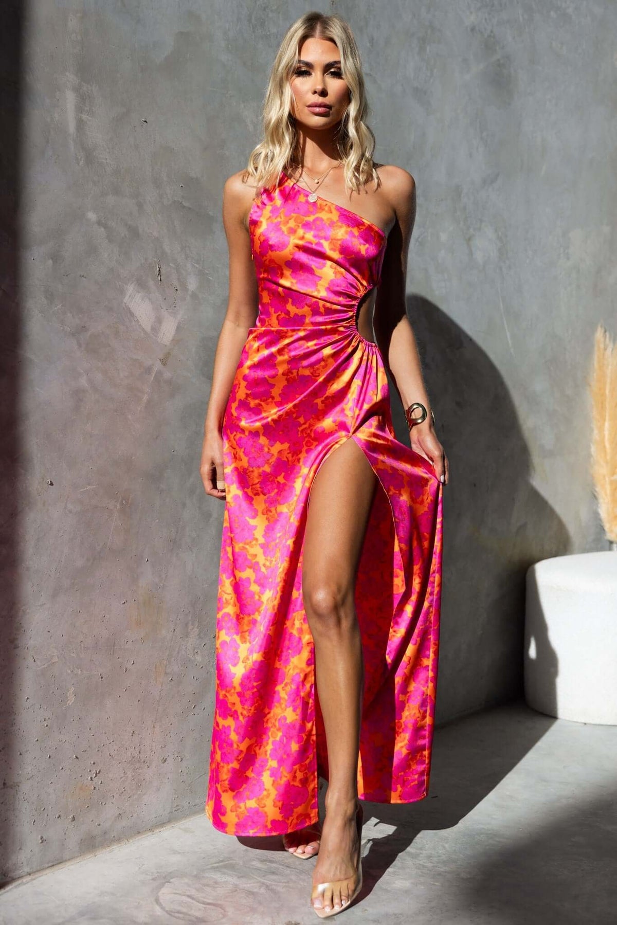 Model wearing Carey Maxi Dress with floral print, one-shoulder neckline, high front split, showcased against a modern backdrop.