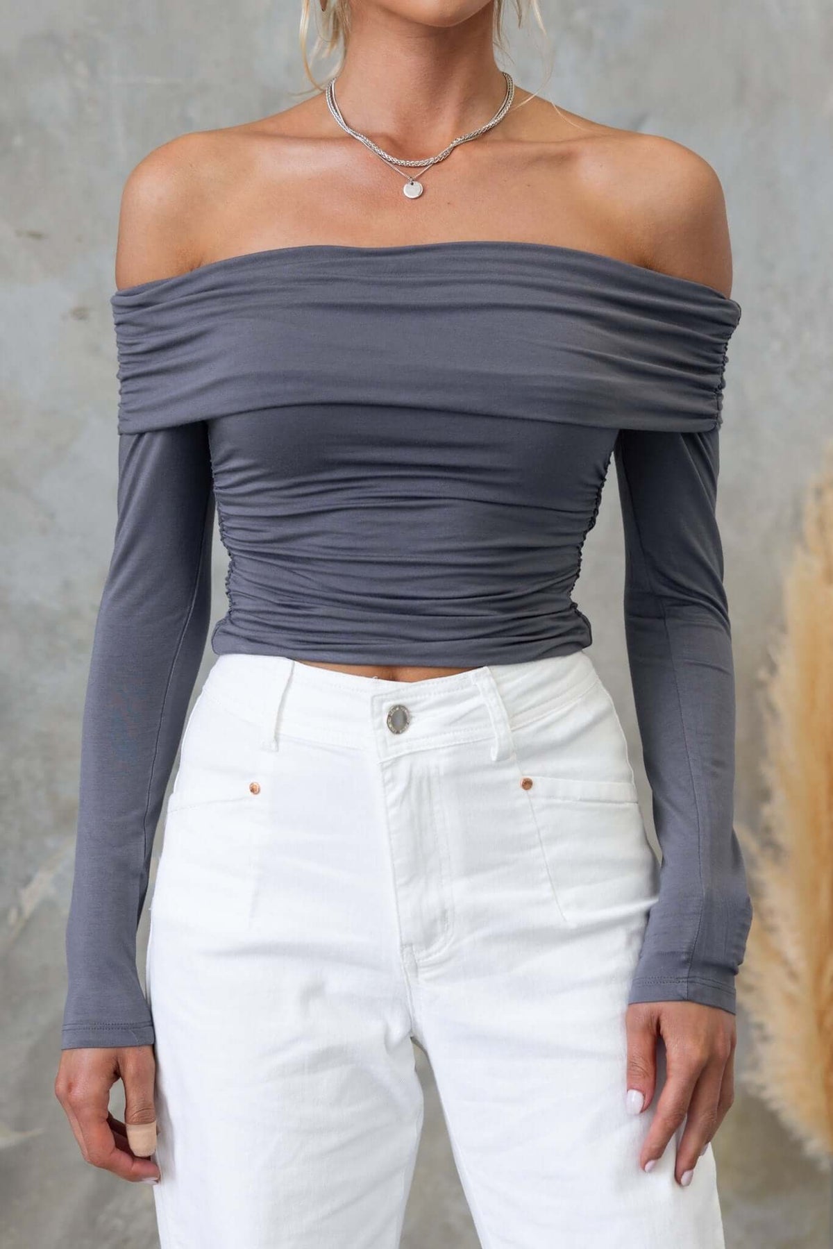 Harrie Top in gray with off-shoulder neckline, long sleeves, and ruched design, paired with white high-waisted jeans.