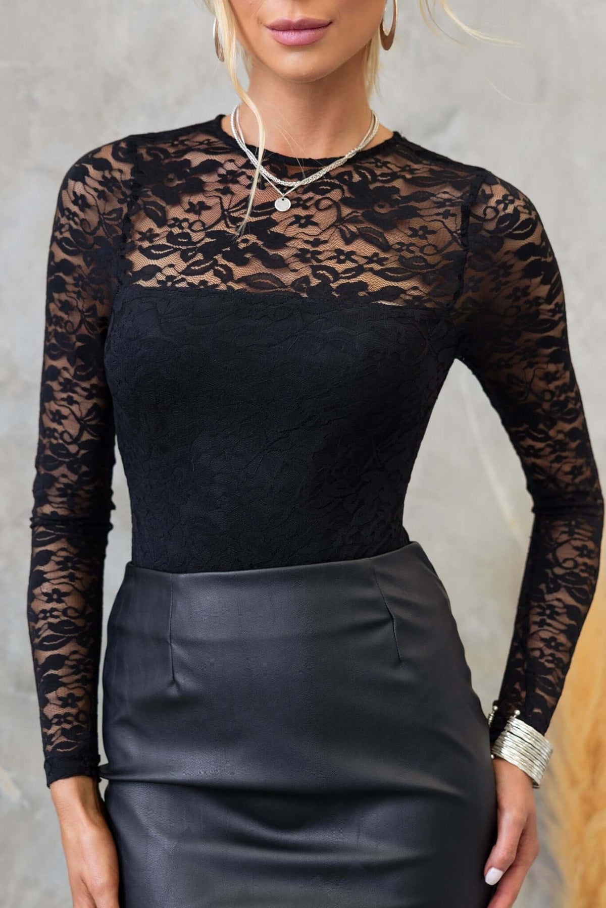 Chic black lace bodysuit with round neckline and long sleeves, styled with a leather skirt and layered jewelry.