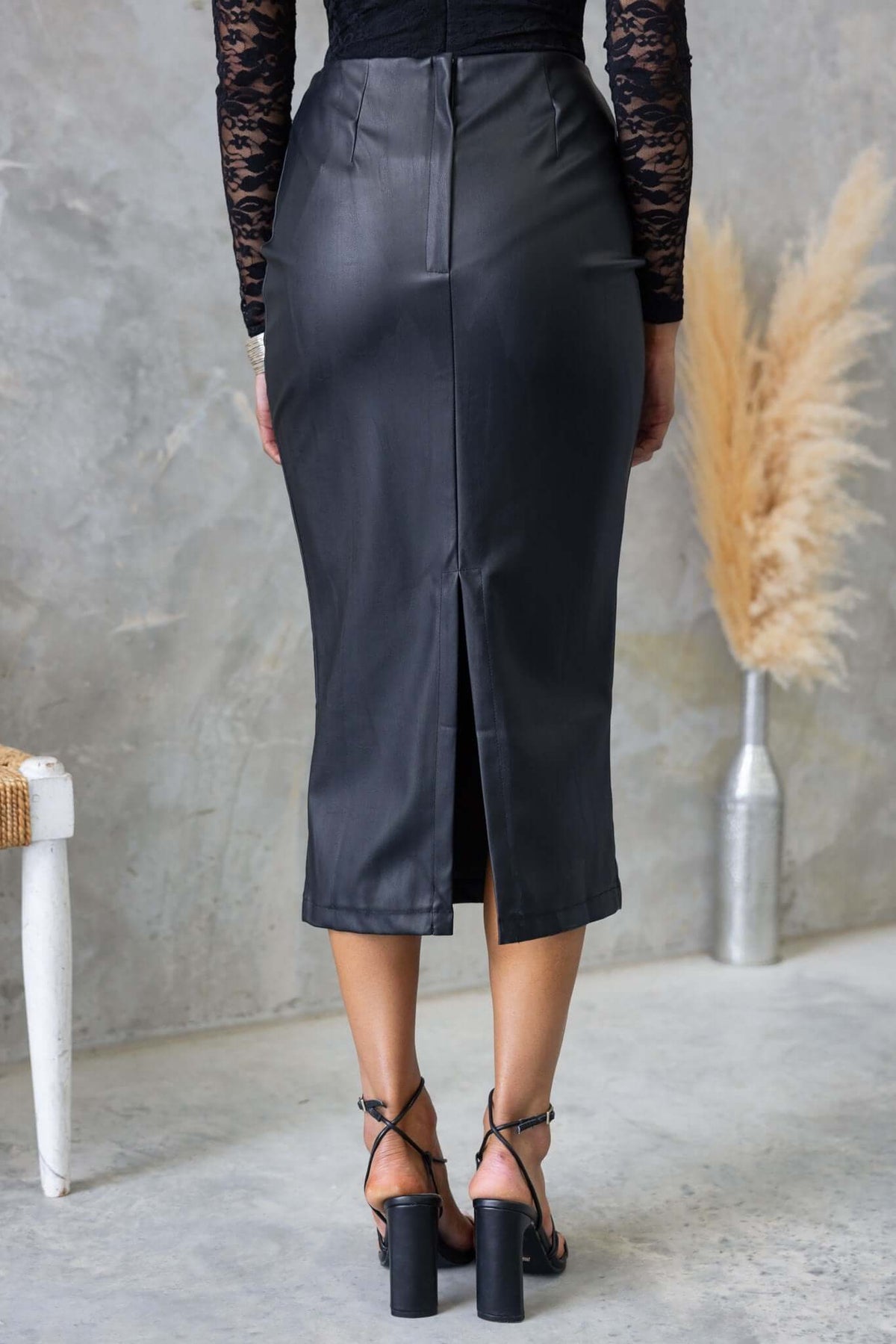 Back view of a black faux leather midi skirt with a split, worn by a model with strappy heels.