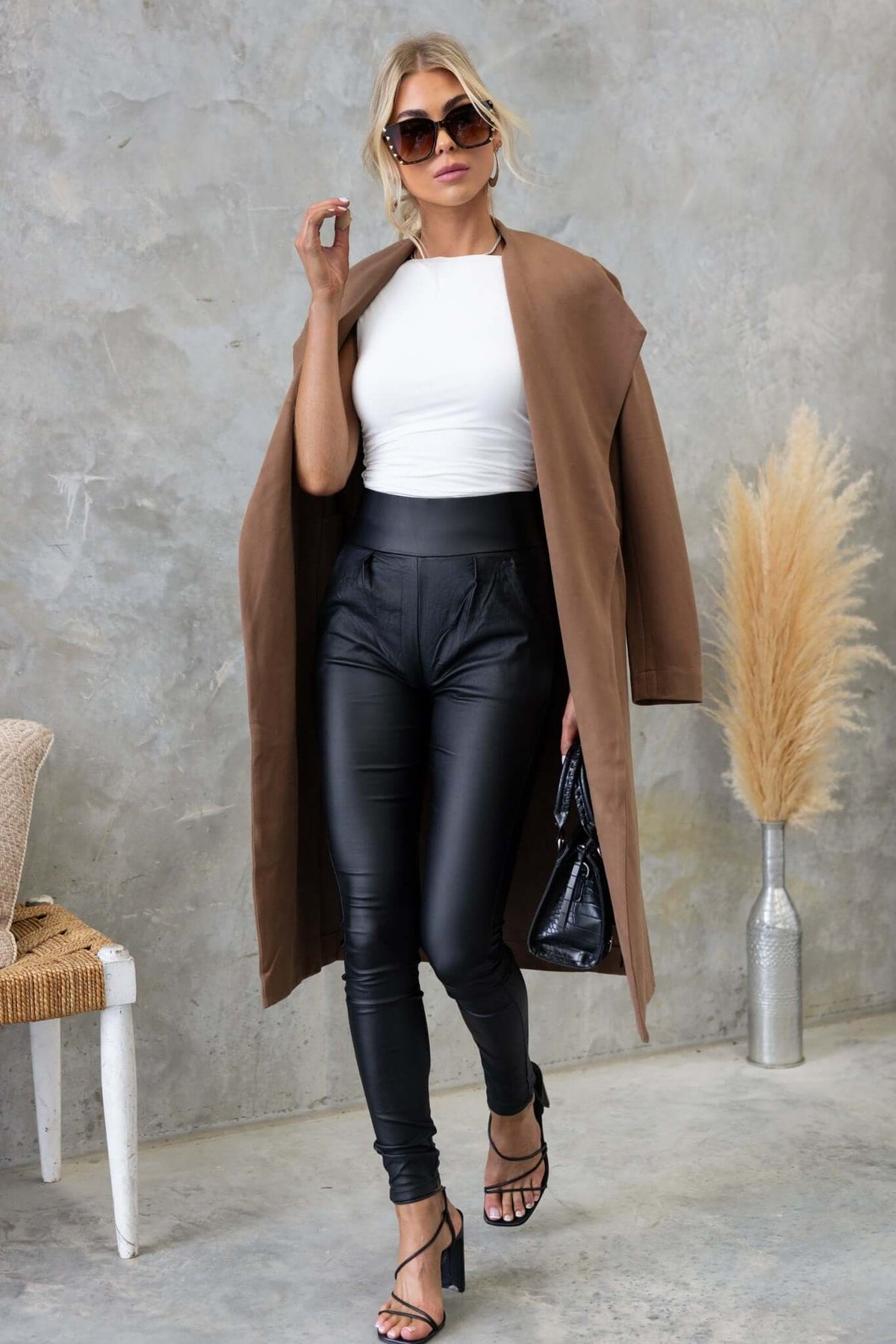 Model wearing the Aria Coat with black faux leather pants and stylish sunglasses in a chic indoor setting.