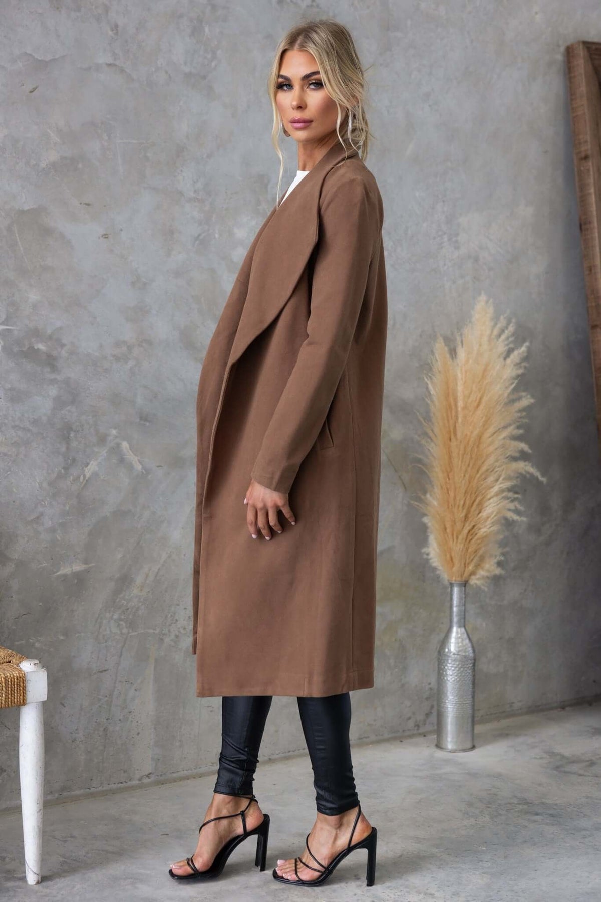 Side view of a woman wearing a longline Aria Coat in brown, styled with black leggings and high heels.