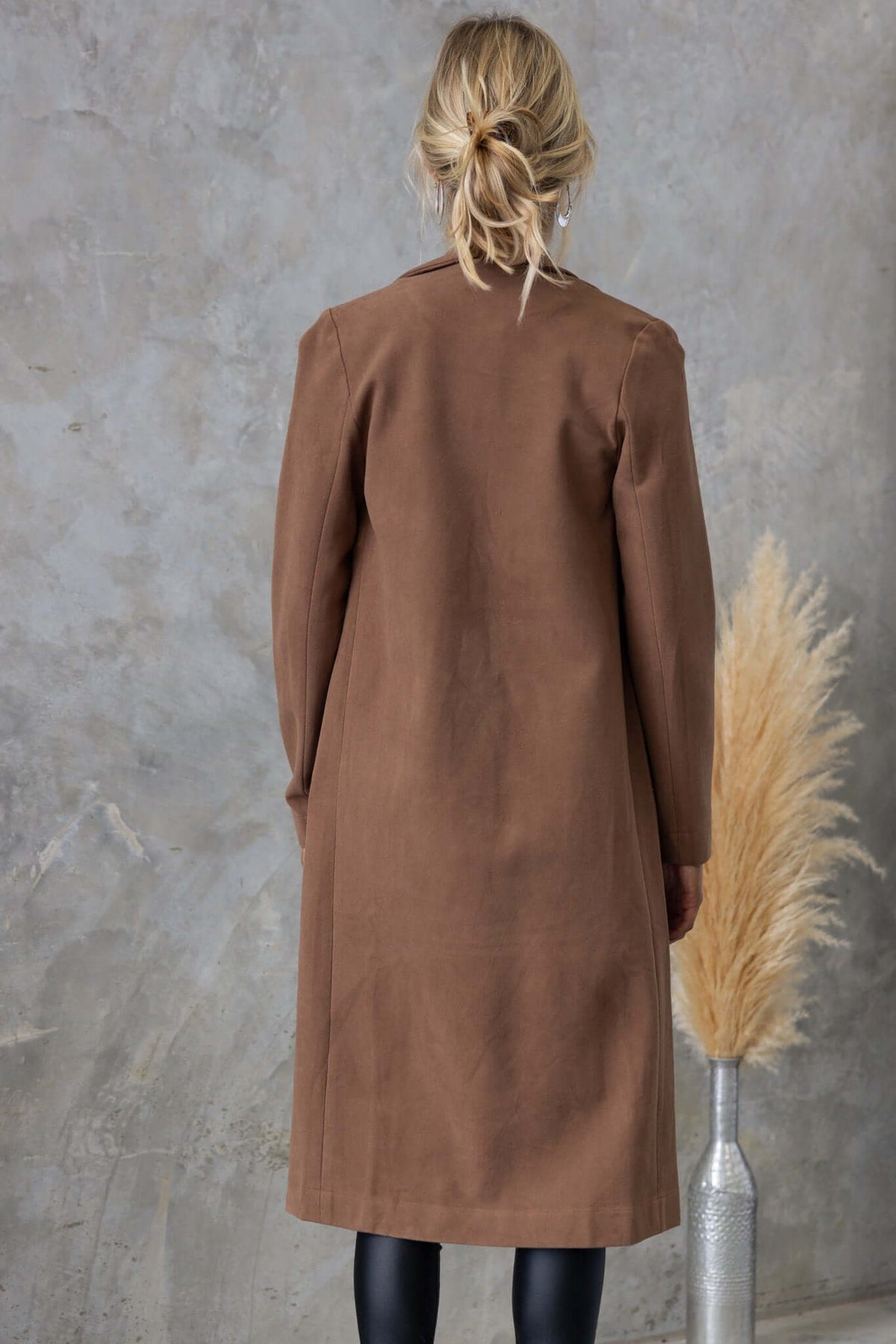 Brown longline Aria coat with collar and lapel, featuring a suede look, styled with a decorative plant.