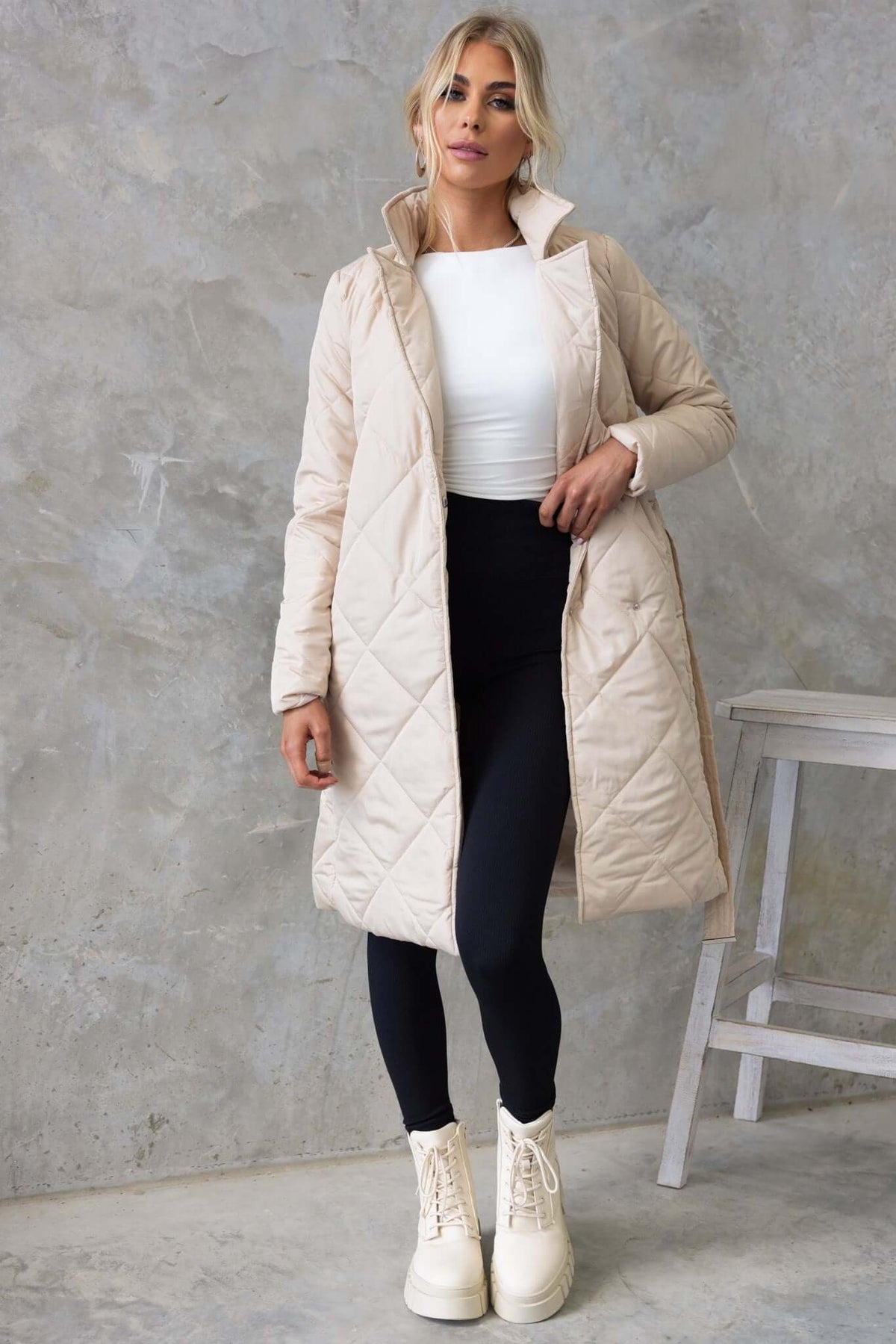 Longline quilted Chantel coat in beige, featuring a collar, lapel, and removable tie, worn with black leggings and boots.